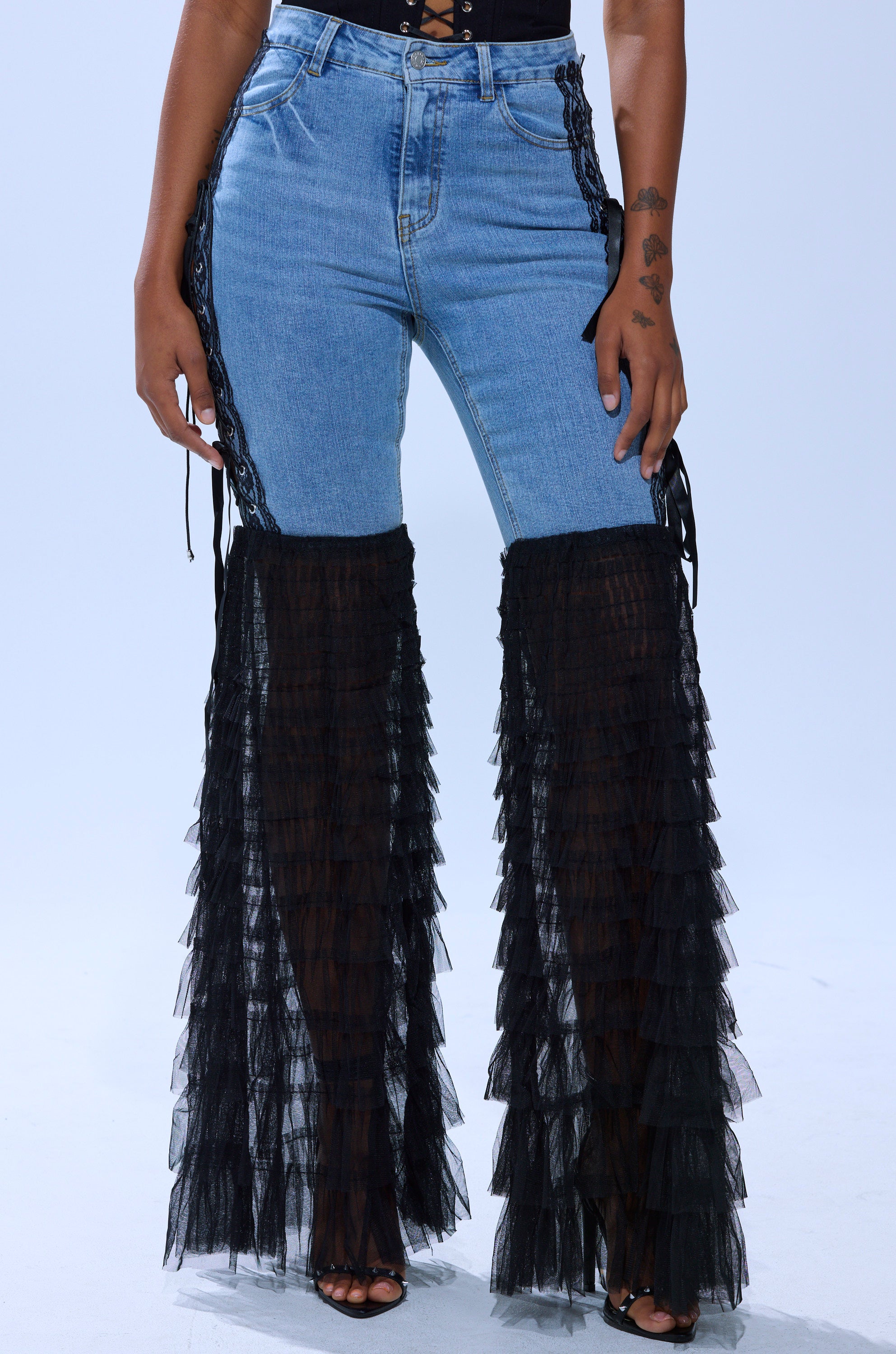 Wearing CAN'T DECIDE TULLE AND LACE DENIM JEANS—high-waisted blue denim with black tulle ruffles from the knees down and lace-up sides—paired with open-toe heels, a person shows small tattoos on their left arm.