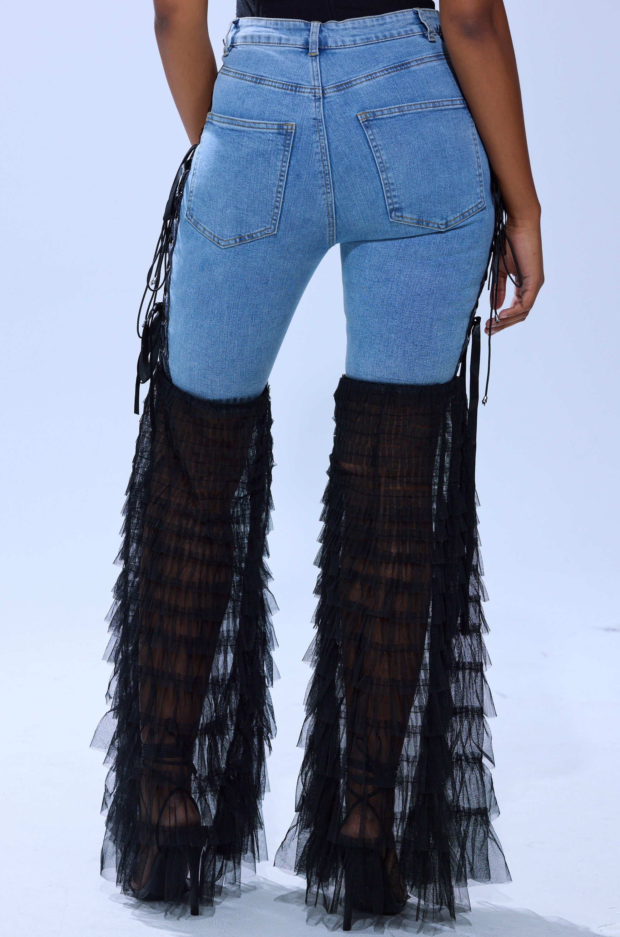 Someone wearing the CAN'T DECIDE TULLE AND LACE DENIM JEANS, featuring sheer black ruffled mesh over denim blue jeans, photographed from the back against a plain light background.