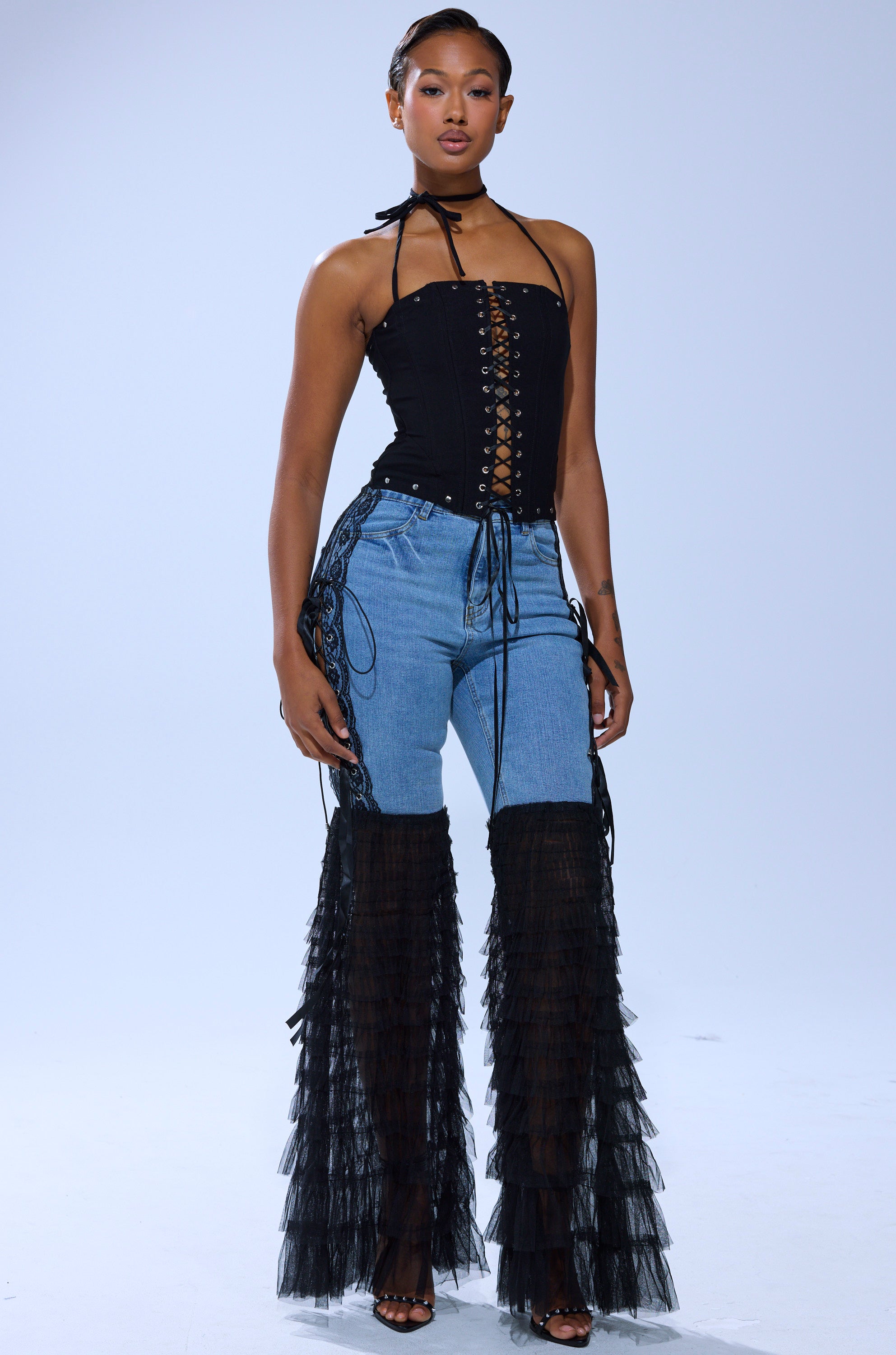 A woman poses in a strapless black lace-up top and CAN'T DECIDE TULLE AND LACE DENIM JEANS featuring dramatic black tulle ruffles from thigh to ankle, paired with black sandals against a plain light background.