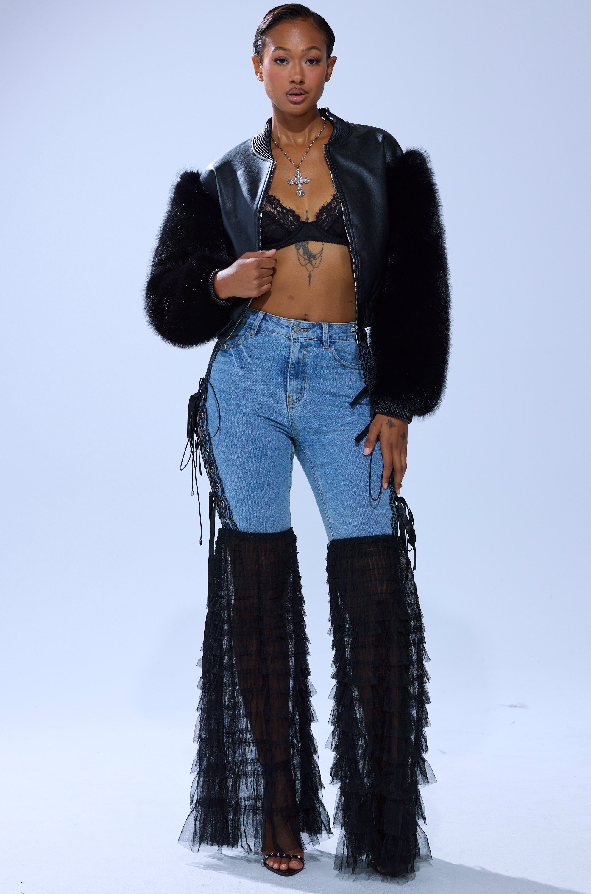 A woman poses confidently in CAN'T DECIDE TULLE AND LACE DENIM JEANS, a black lace bralette, fur-accented jacket, sheer ruffled leg coverings, layered necklaces, and open-toe heels against a plain backdrop.