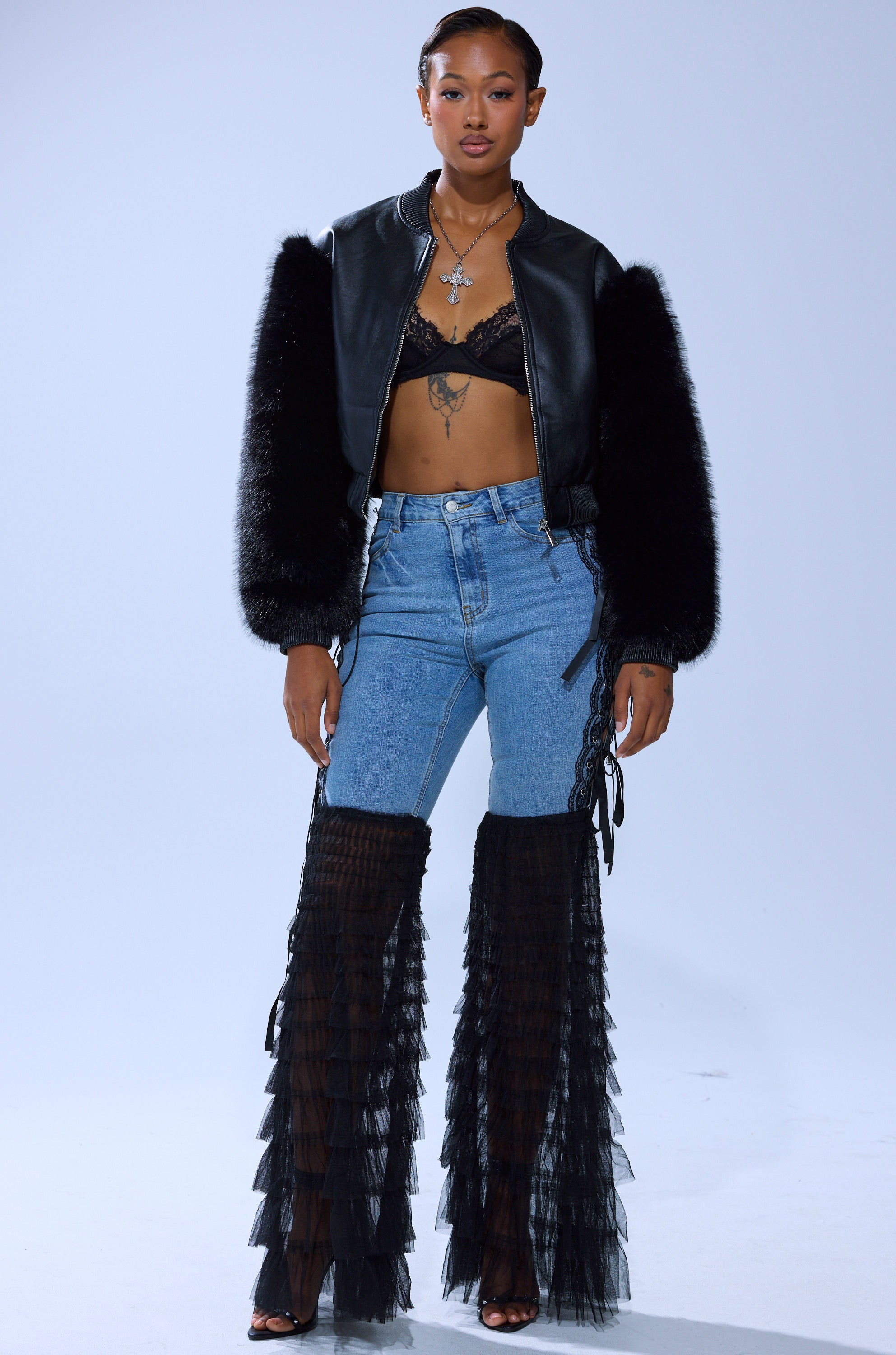 A woman with short hair and layered necklaces stands confidently in CAN'T DECIDE TULLE AND LACE DENIM JEANS, styled with a black fur-sleeved jacket, lace bralette, sheer tiered ruffle chaps, and open-toed heels.