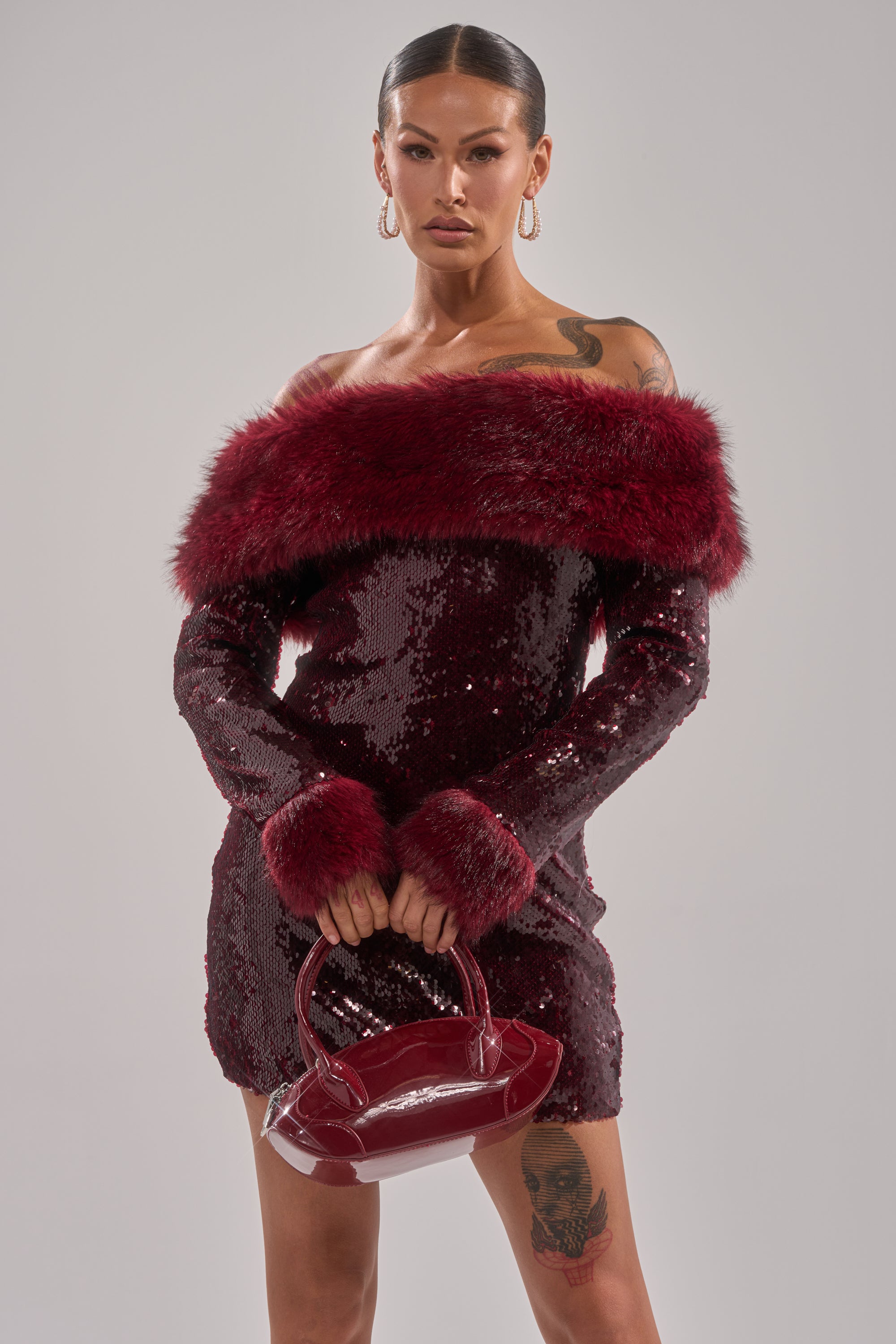 A woman wears the BABY IT'S COLD OUTSIDE FUR TRIM SEQUIN MINI DRESS in dark red with faux fur trim, holding a shiny red handbag. She accessorizes with hoop earrings, has slicked-back hair, and tattoos on her arm and thigh.
