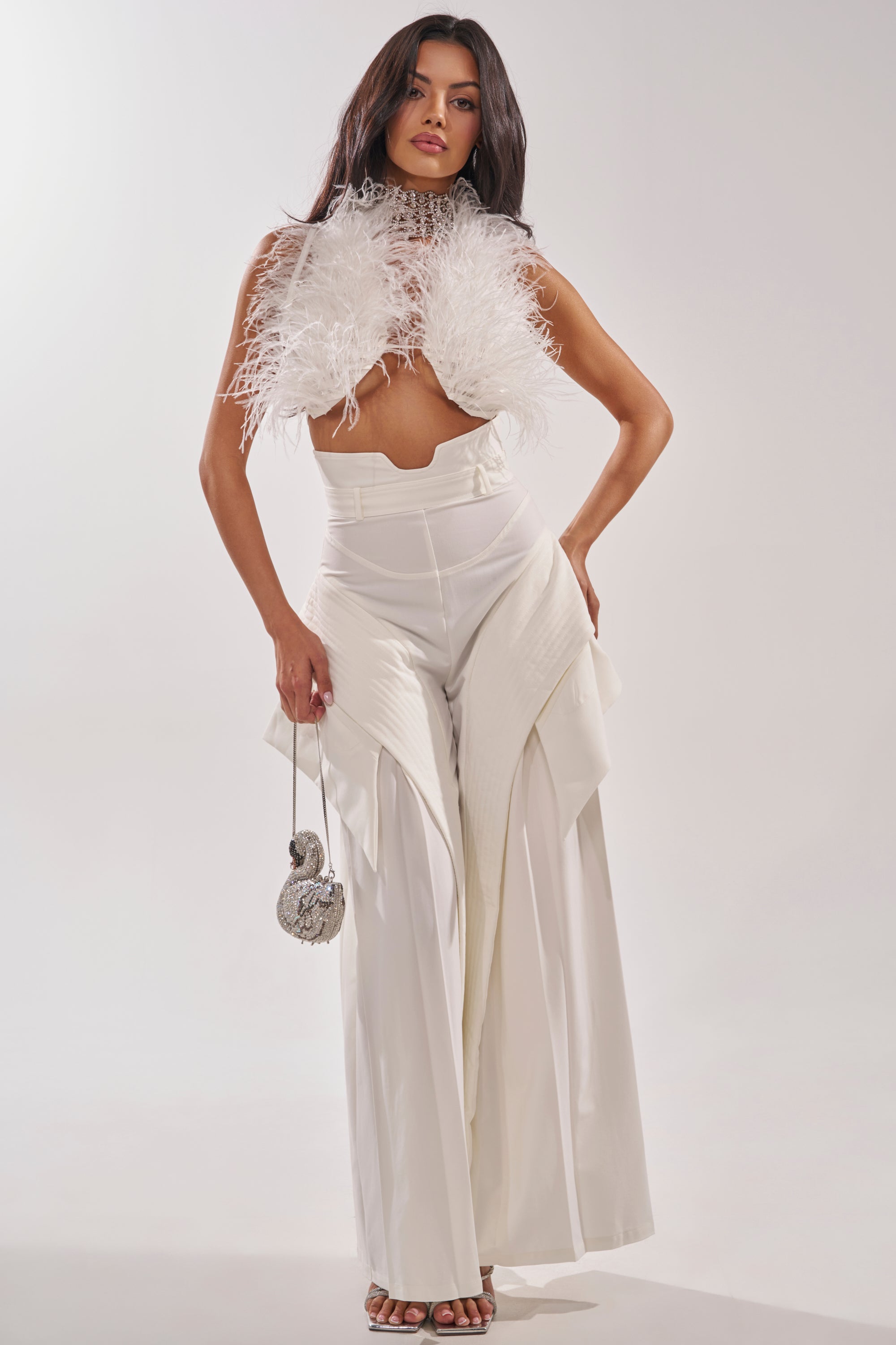 A woman poses confidently in luxury streetwear—a feathered crop top with the TOO COOL FOR YOU TROUSER. She holds a small silver round purse and wears open-toed heels, standing out against a light background. 0
