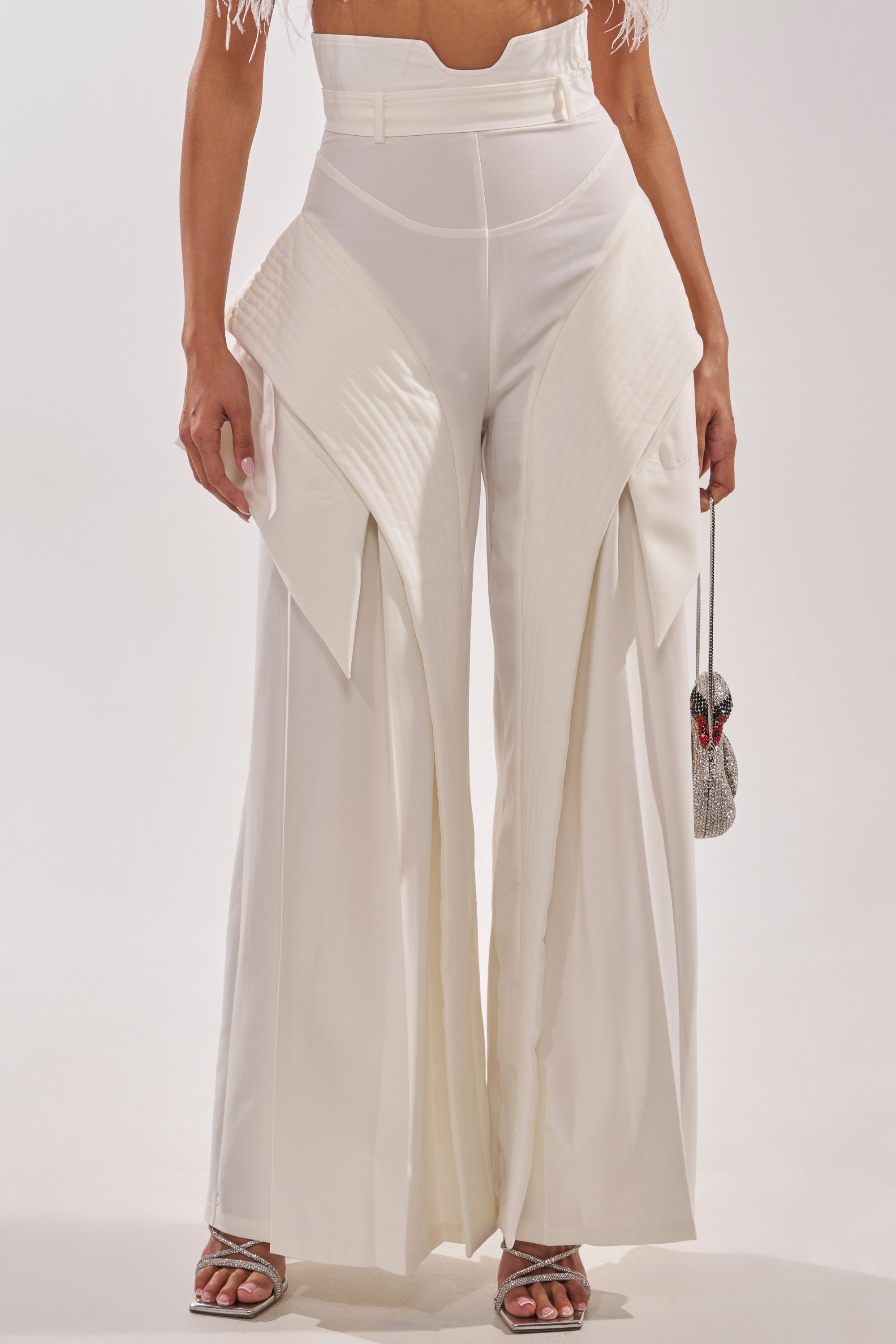 A person wears the TOO COOL FOR YOU TROUSER—high-rise, wide-leg with layered, angular panels—paired with silver strappy heels and a sparkly silver mini handbag, set against a plain white background for a luxe streetwear vibe. 1