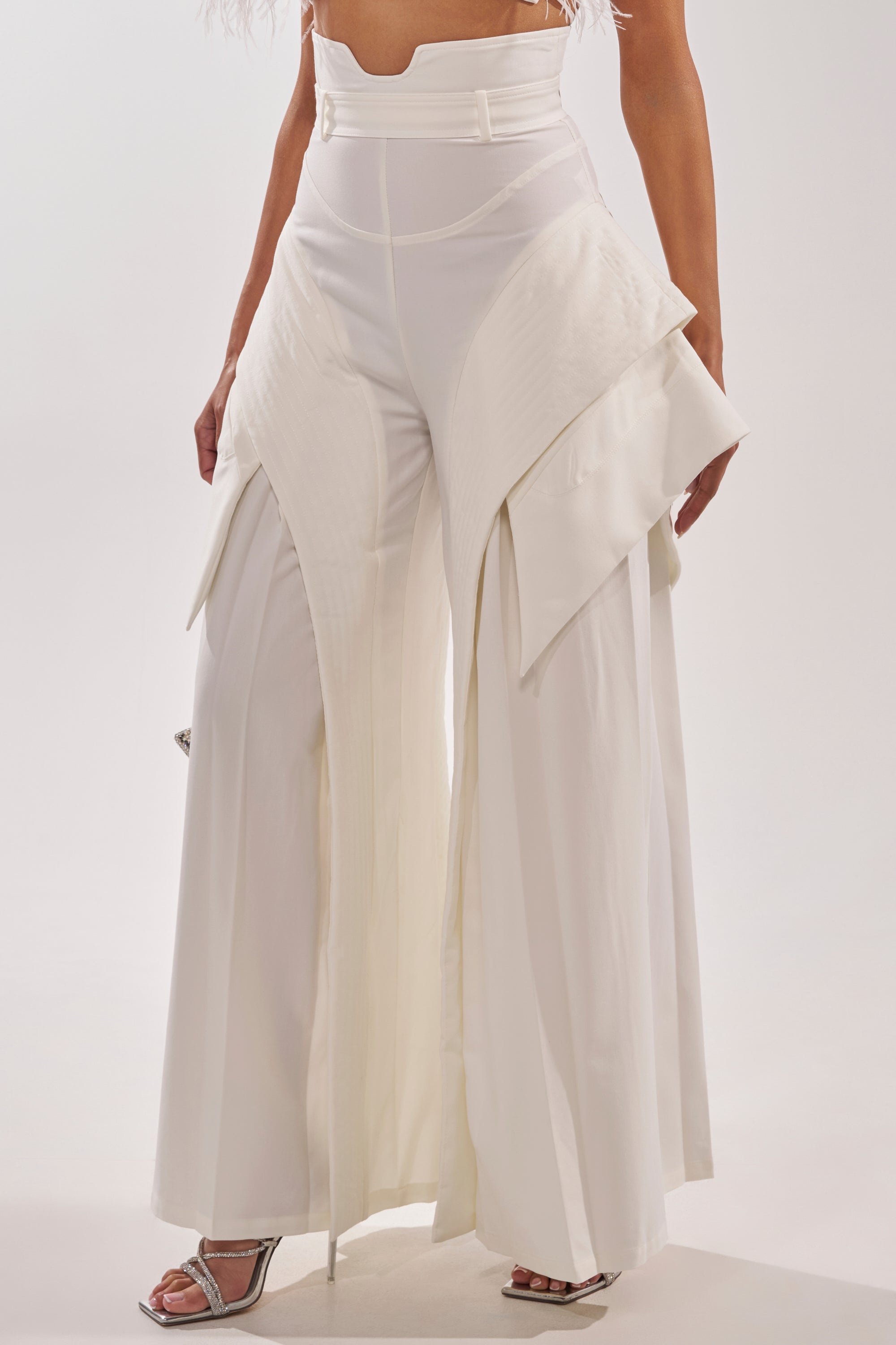 Wearing the TOO COOL FOR YOU TROUSER—high-rise, wide-leg white pants with sculptural layers and a geometric design—paired with silver open-toe heels for a luxury streetwear vibe. Only the lower body is visible. 3