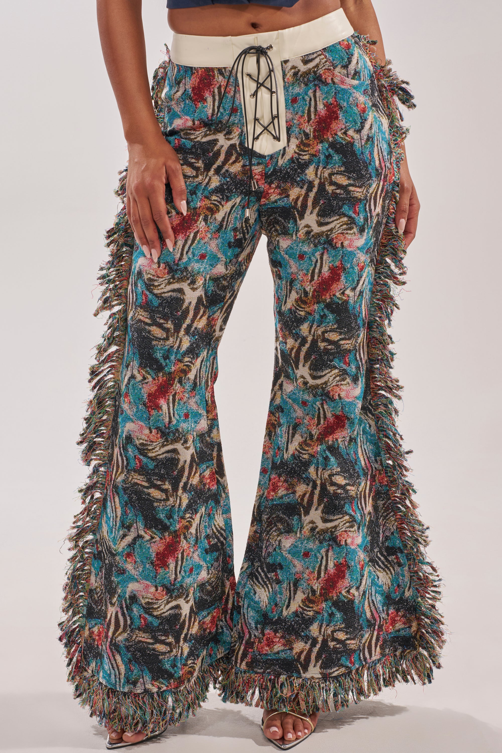 A person wears the MIXED EMOTIONS TAPESTRY PANT, featuring a multicolor abstract print in blue, red, and beige, fringe side trim, a lace-up white waistband, and flared legs for a textured bohemian style.