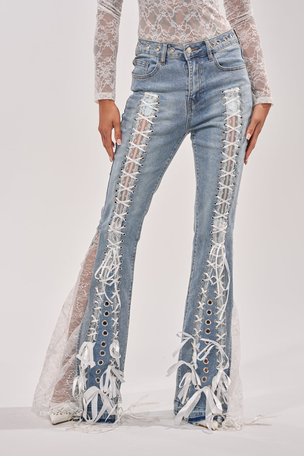 A person wears the LOVE ME LIKE THAT LACE UP FLARE DENIM—light blue high-rise wide-leg jeans with sheer white lace panels, white ribbon and grommet details—paired with a long-sleeve white lace top. 0