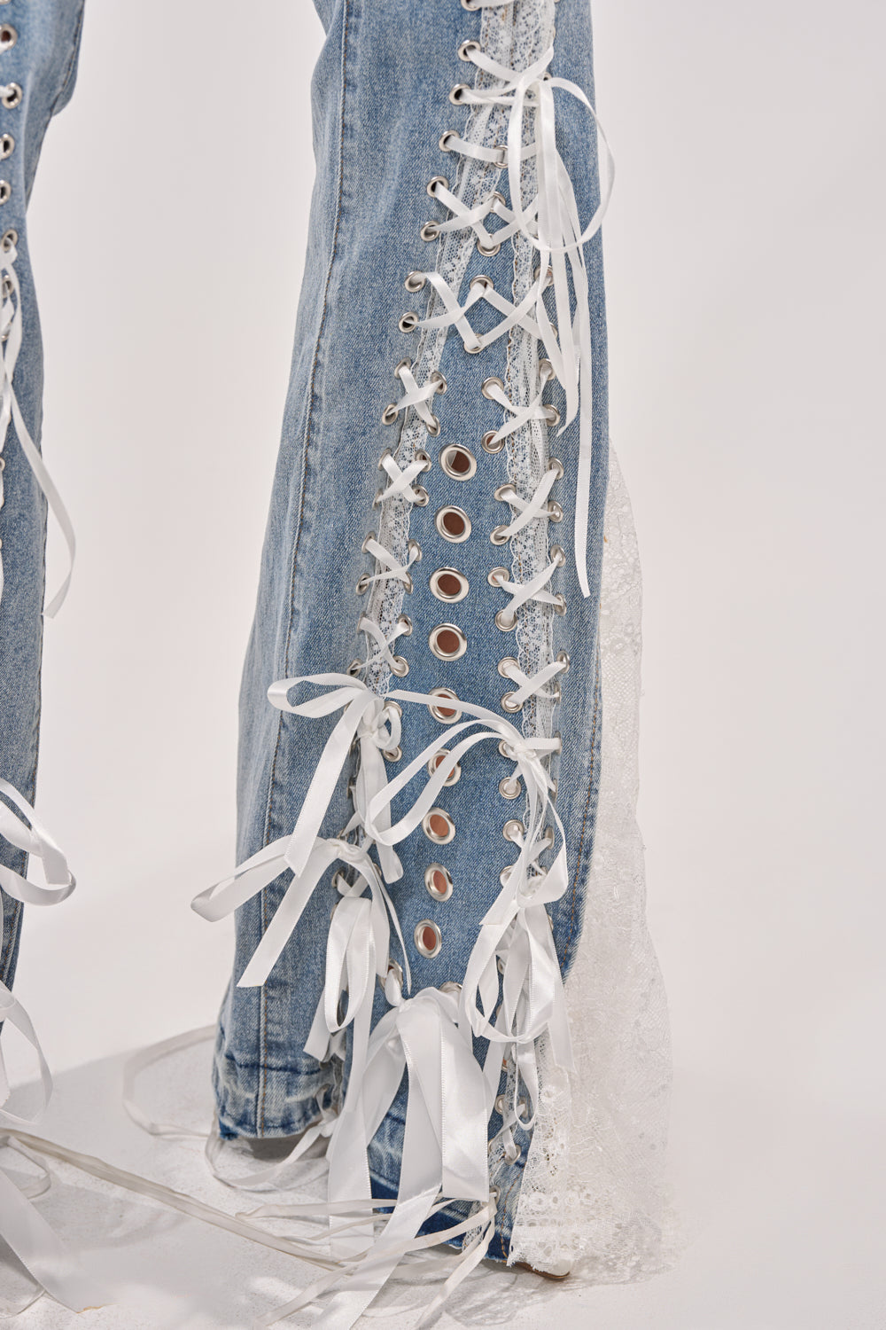 Close-up of LOVE ME LIKE THAT LACE UP FLARE DENIM featuring statement flared legs, large white eyelets, ribbon laces tied in bows along the side seam, and a hint of white lace fabric peeking out near the hem. 9