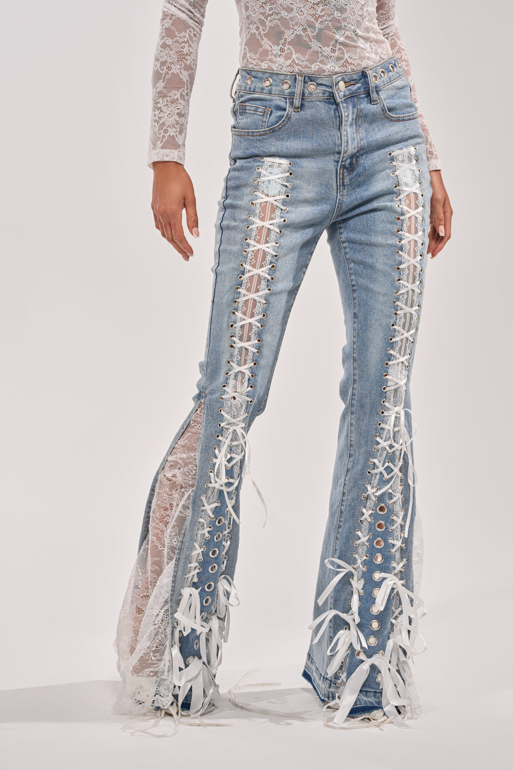A person wears the LOVE ME LIKE THAT LACE UP FLARE DENIM—light blue, high-rise flared jeans with white lace-up front panels and ribbon-trimmed hems—paired with a long-sleeve white lace top. Head and feet are not visible. 1