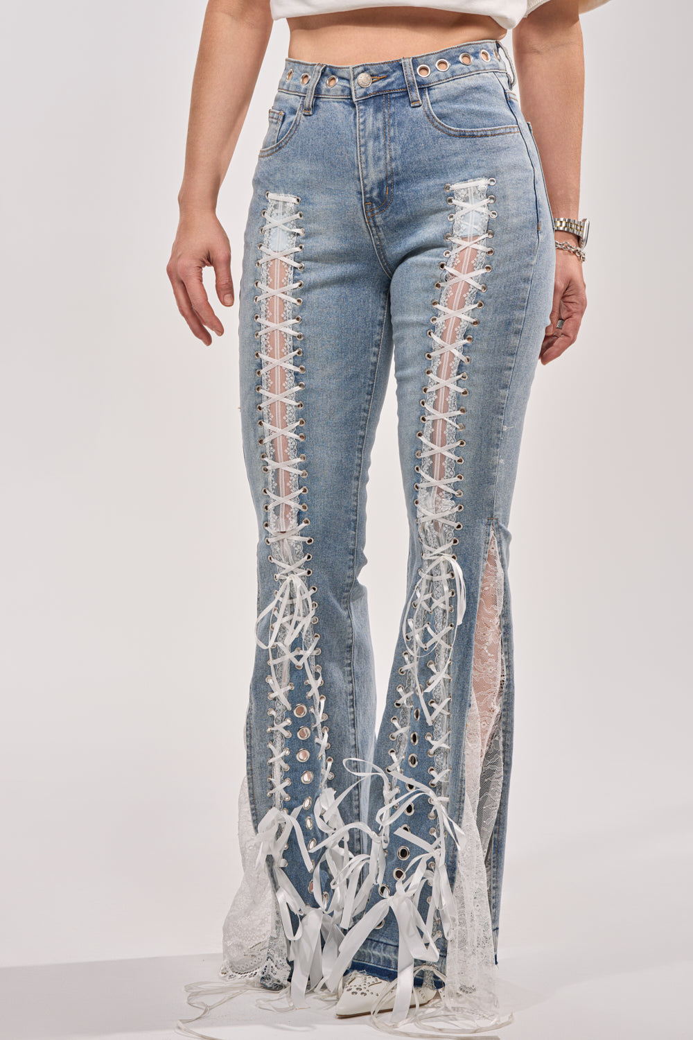 A person wears the LOVE ME LIKE THAT LACE UP FLARE DENIM—high-rise blue jeans with lace-up panels, sheer lace details, frayed hems, and decorative white bows at the ankles—paired with a white crop top. 2