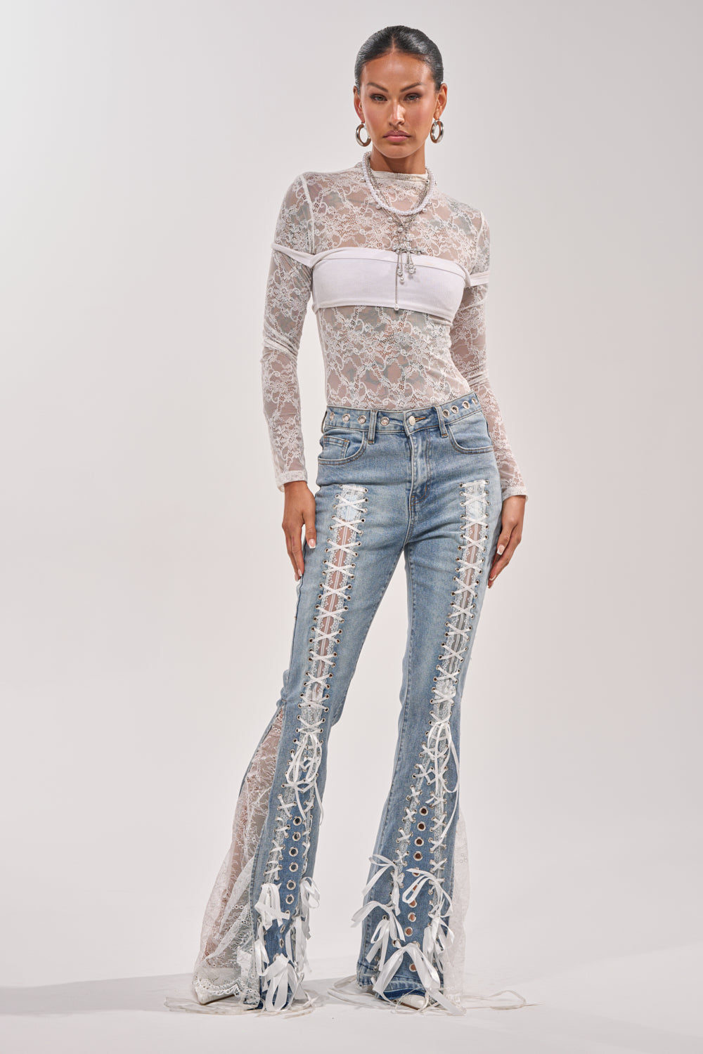A woman models the LOVE ME LIKE THAT LACE UP FLARE DENIM—blue, high-rise flared jeans with lace-up slits and front tassel details—paired with a sheer white lace top over a bandeau, hoop earrings, and hair pulled back. 6