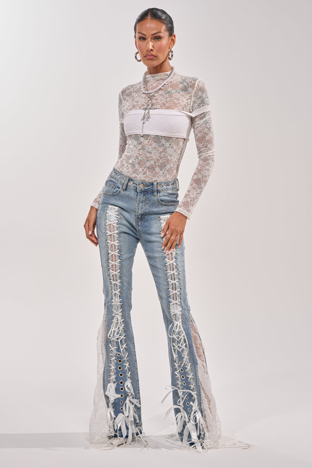 A woman poses against a plain background wearing blue LOVE ME LIKE THAT LACE UP FLARE DENIM with bold front lace-up panels and white ribbon ties, styled with a sheer white lace top over a white bandeau. 7