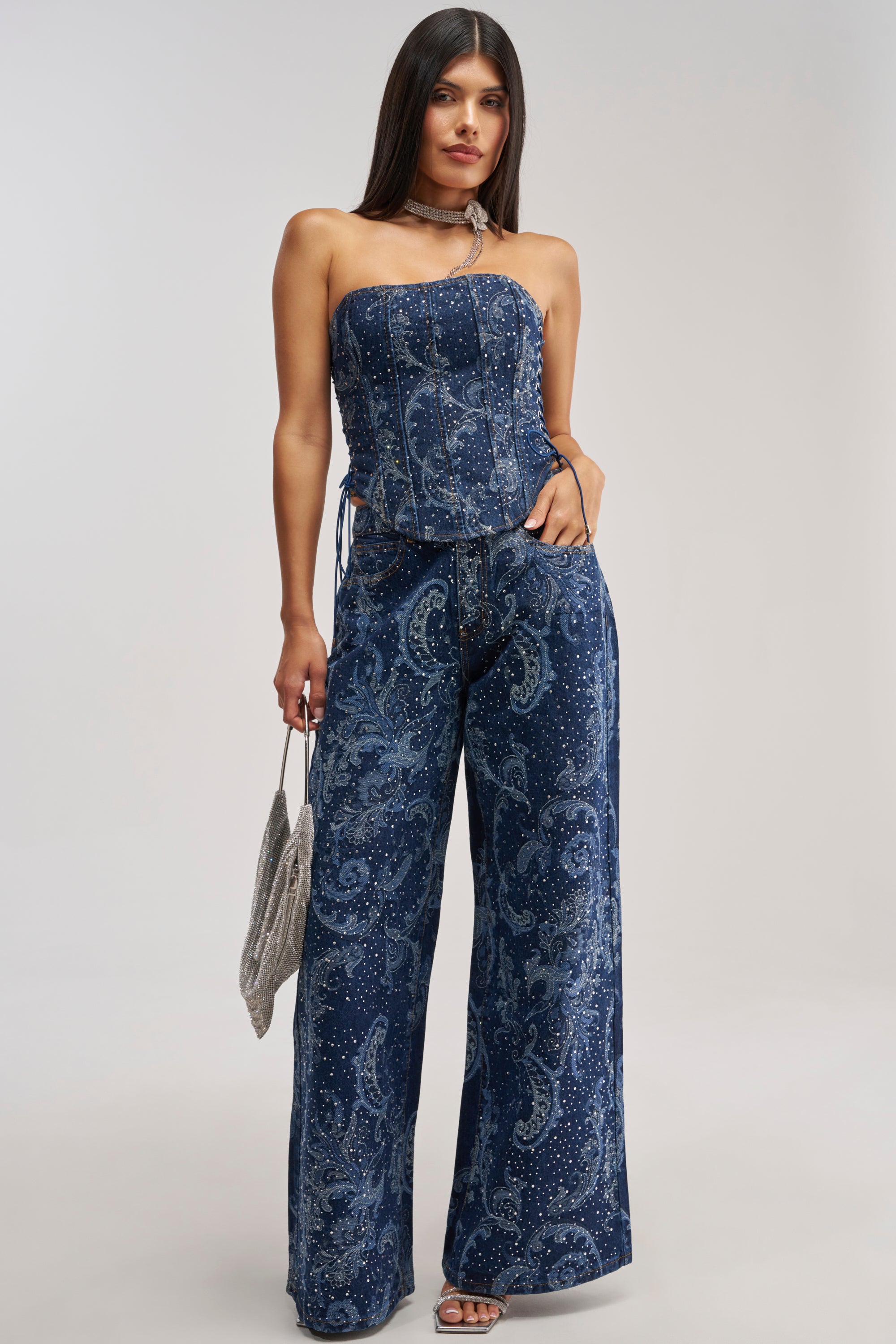 A woman wearing a strapless blue paisley corset top and the ELEVATED WIDE LEG DENIM WITH RHINESTONES, which features a flattering high-rise waist, holds a silver bag while standing against a plain light background.