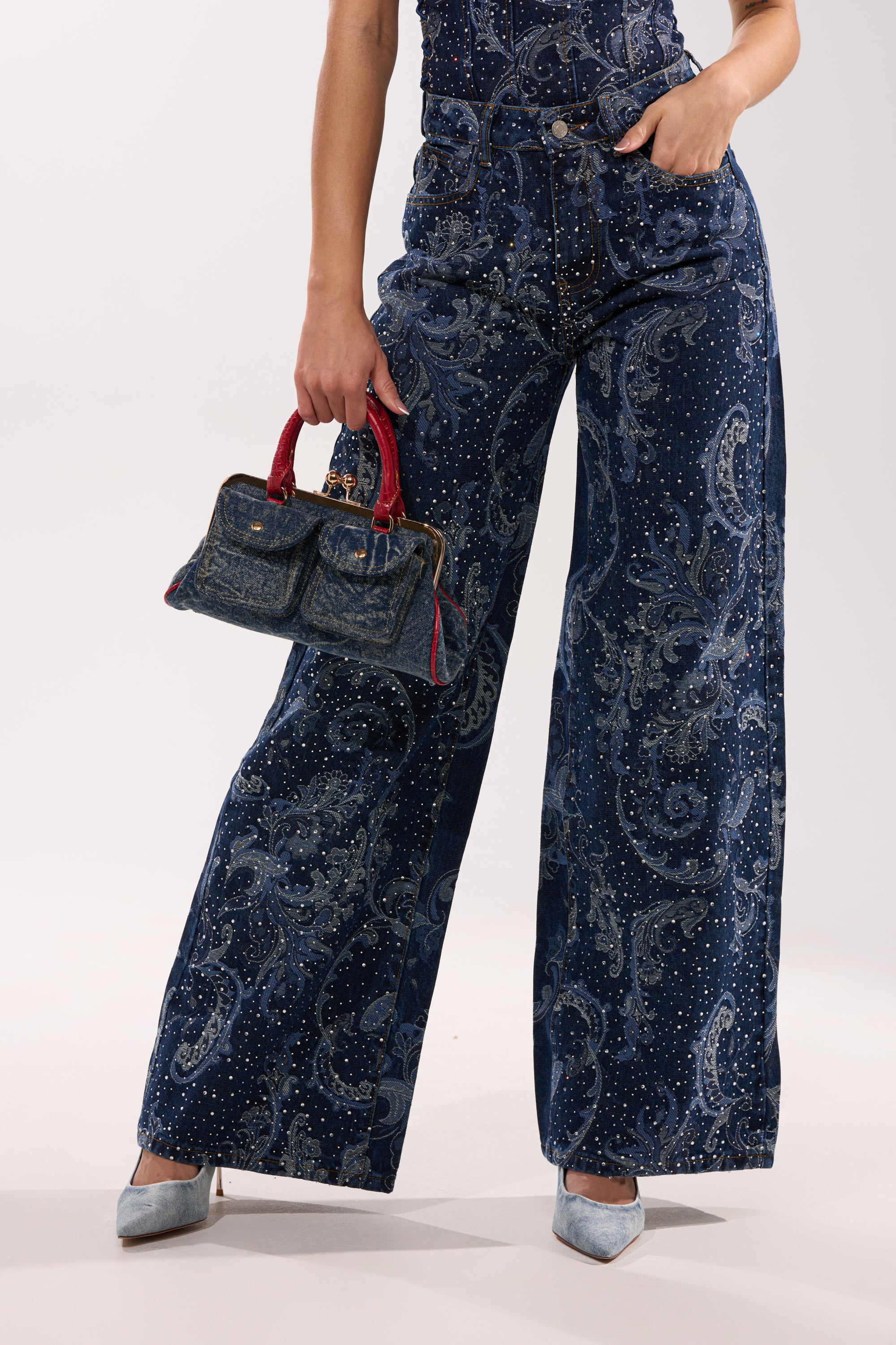 Wearing the ELEVATED WIDE LEG DENIM WITH RHINESTONES, a person holds a denim handbag with red handles. Only the lower half is shown, featuring light blue pointed-toe heels and sparkling denim details.