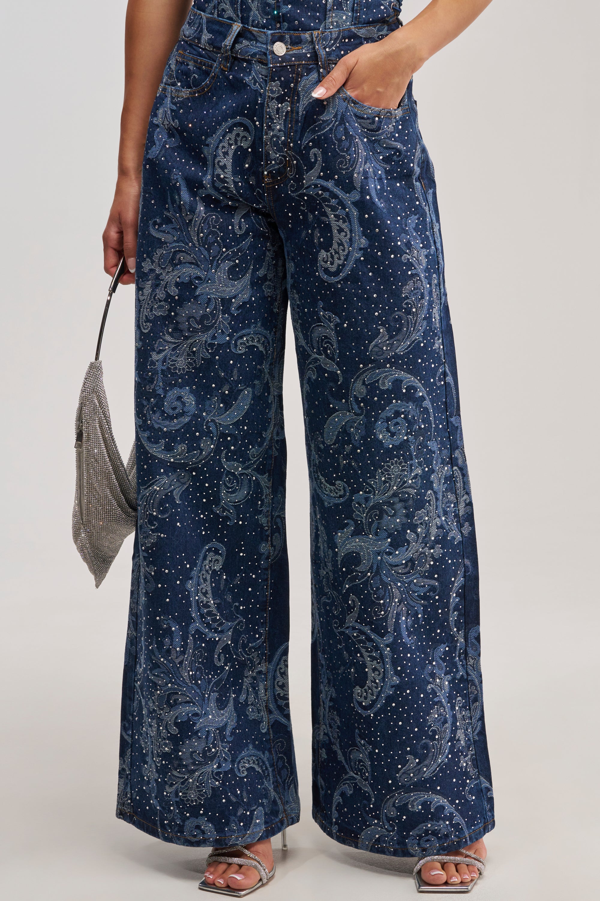 A person wearing the ELEVATED WIDE LEG DENIM WITH RHINESTONES—high-rise, wide-leg jeans adorned with paisley rhinestone embellishments in dark blue tones—paired with silver open-toe heels and a small silver handbag. The image is cropped from shoulder to feet.