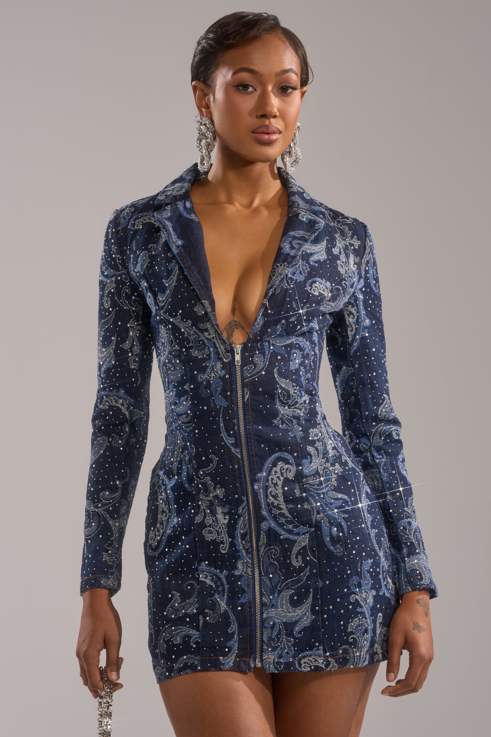 Woman wears the INTO SOMETHING BETTER DENIM BLAZER MINI DRESS, a fitted deep blue mini dress with rhinestone details, zipper front, and paisley patterns. She pairs it with statement earrings and styled hair against a plain background.