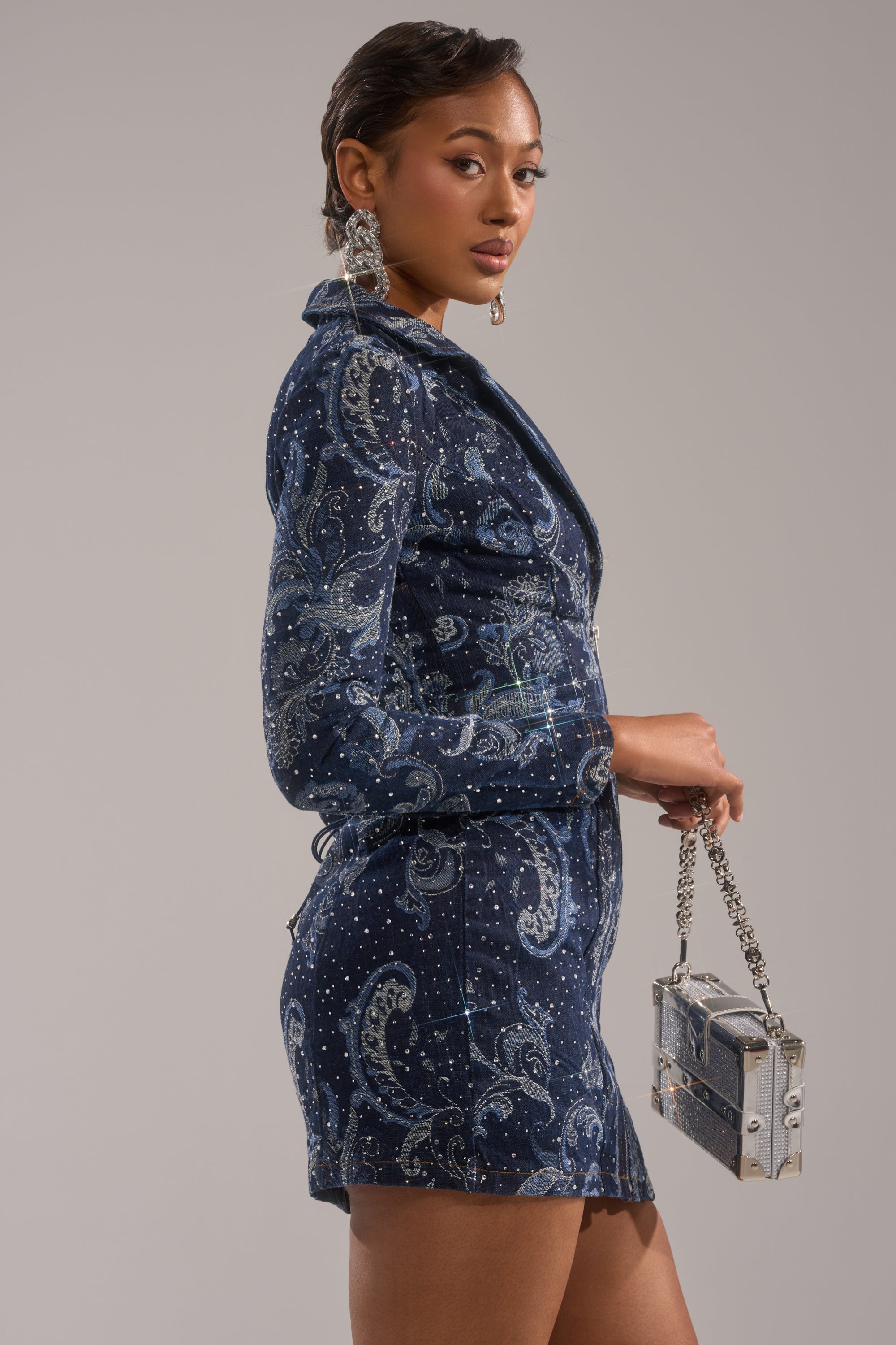 A woman wears the INTO SOMETHING BETTER DENIM BLAZER MINI DRESS in blue paisley print with rhinestone details, paired with a matching chain-strap handbag. She has short hair and statement earrings, standing against a plain gray background.