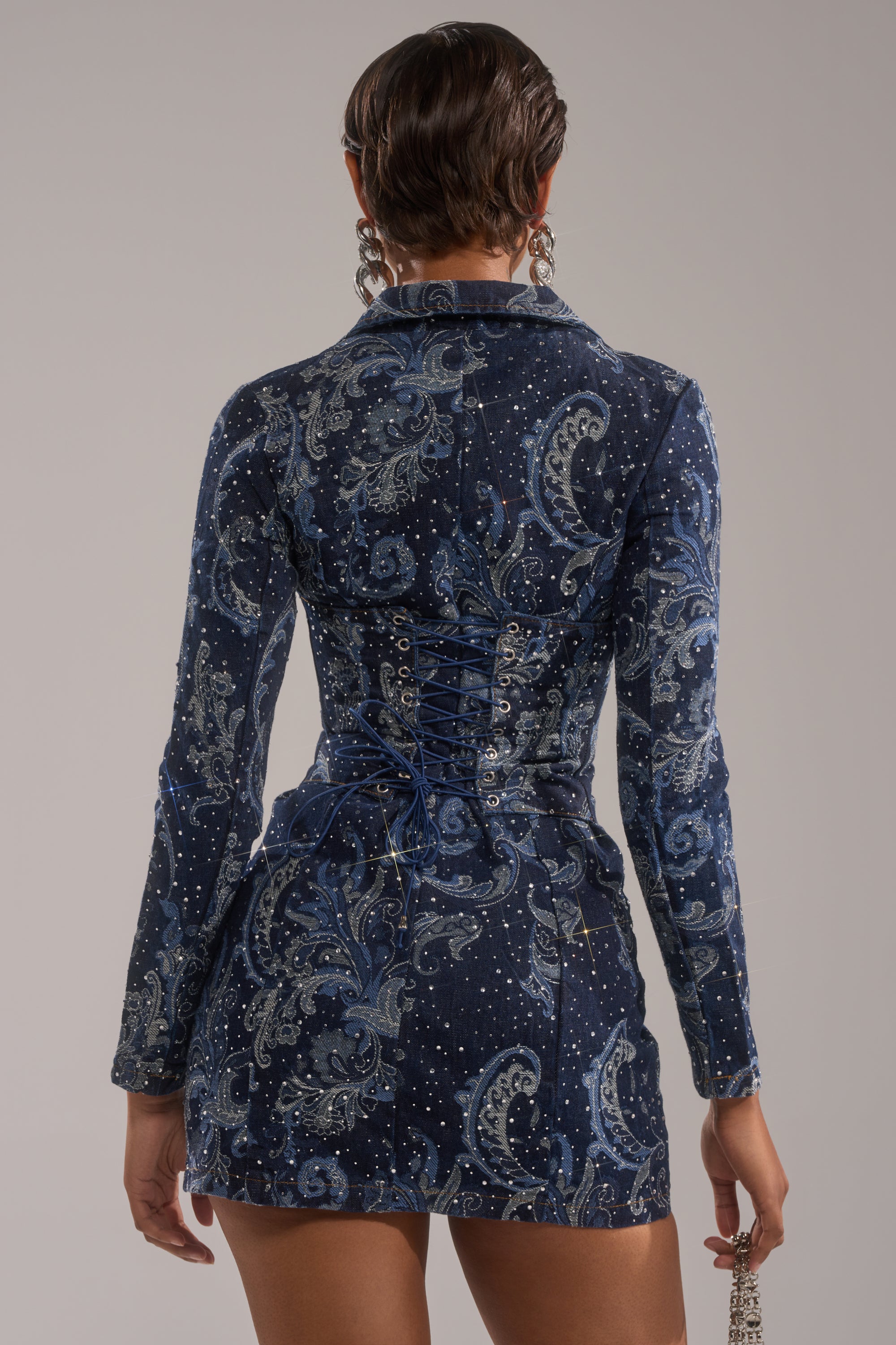 A person with short, slicked-back hair wears the INTO SOMETHING BETTER DENIM BLAZER MINI DRESS, seen from behind against a plain background.