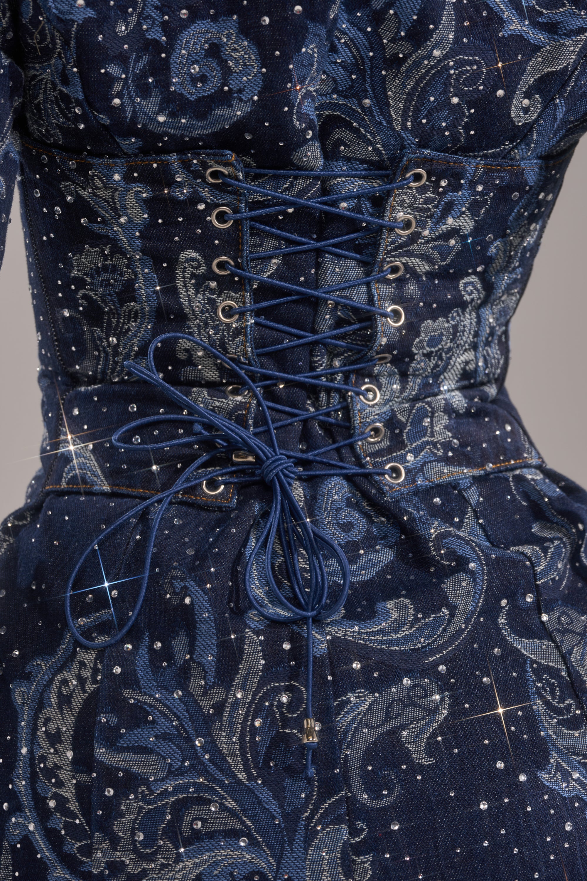 Close-up of the INTO SOMETHING BETTER DENIM BLAZER MINI DRESS featuring a dark blue fitted corset design with silver paisley print, sparkling rhinestones, and blue crisscross laces at the back.