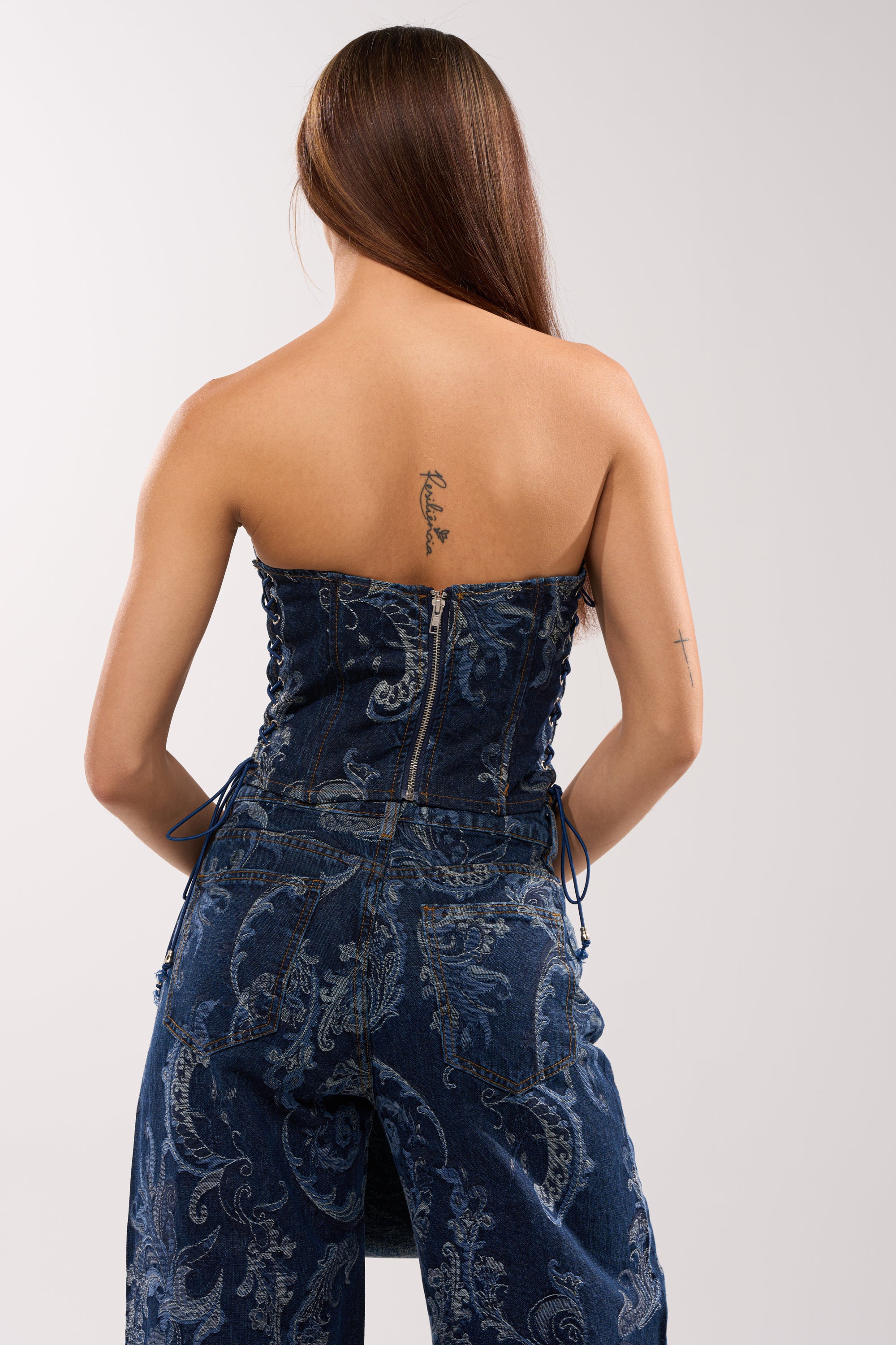A woman with long brown hair, seen from behind, wears the ELEVATED DENIM CORSET WITH RHINESTONES and matching jeans. Tattoos on her upper back and right arm complete her standout festival style. 3