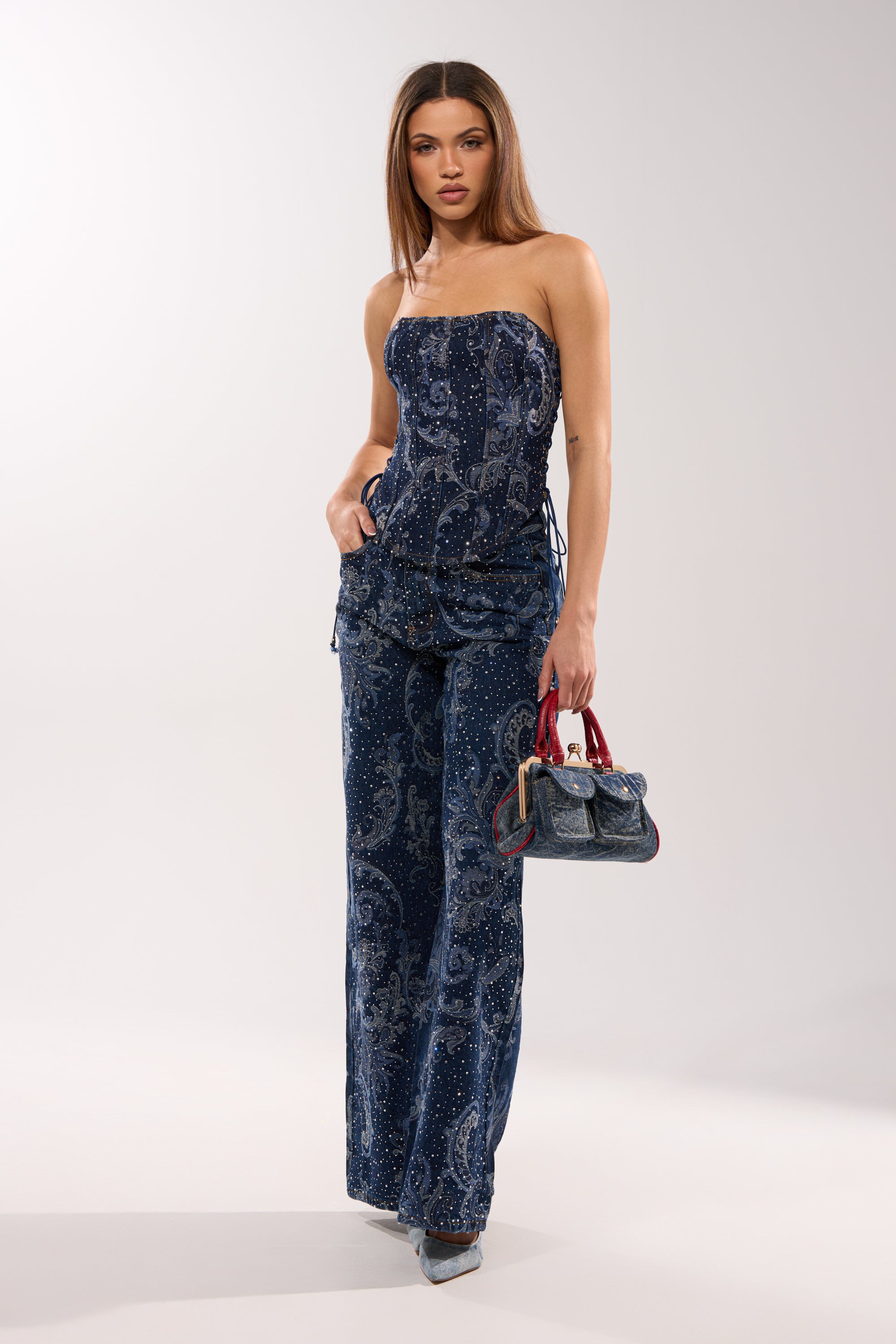 A woman wears the ELEVATED DENIM CORSET WITH RHINESTONES jumpsuit, paired with silver pointed heels, and holds a small blue handbag with red handles against a plain light background. 5