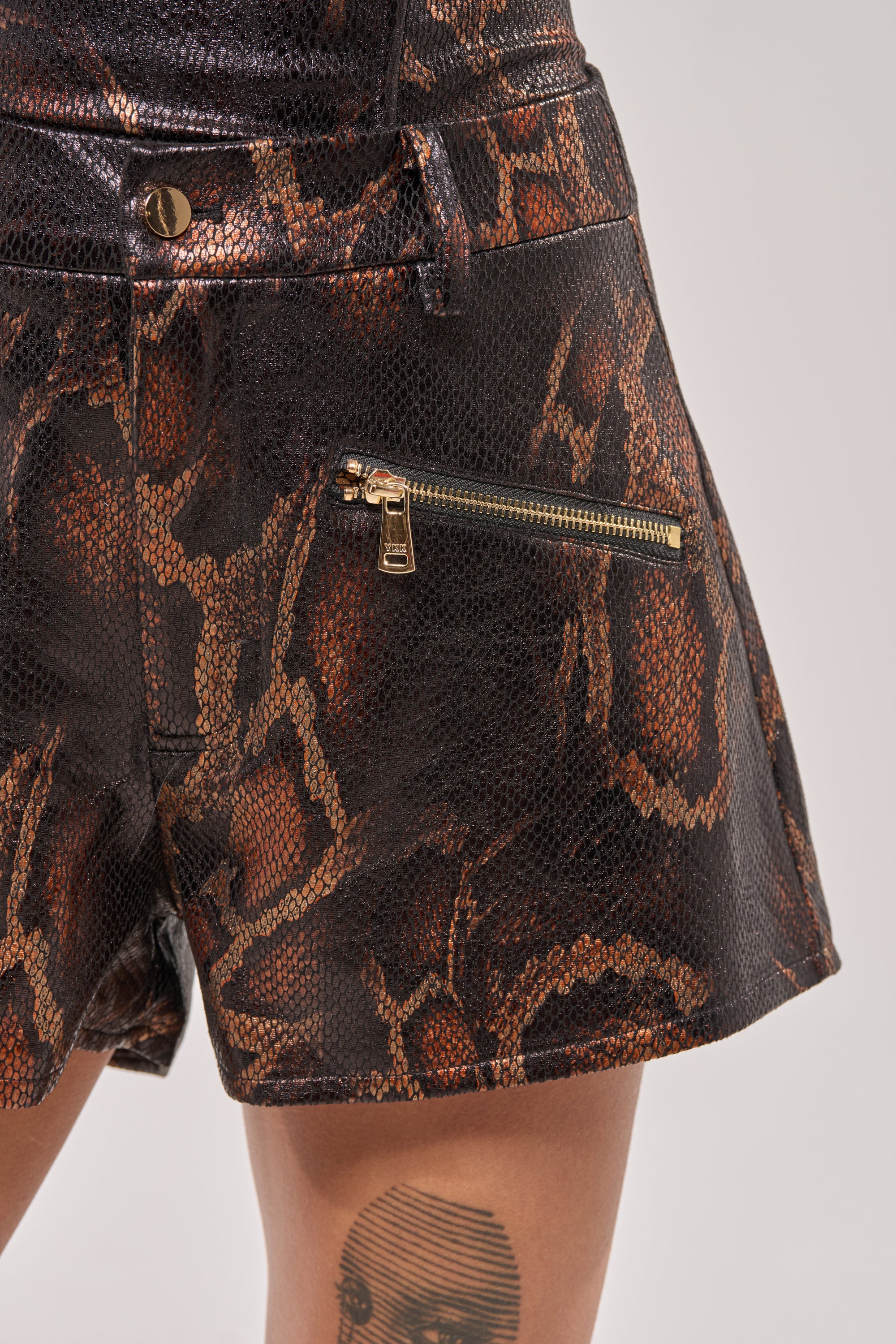 Close-up of a person wearing BEING BAD SNAKE SHORTS featuring a brown snake print and gold zipper pocket. A black circular tattoo is visible on their thigh. 8