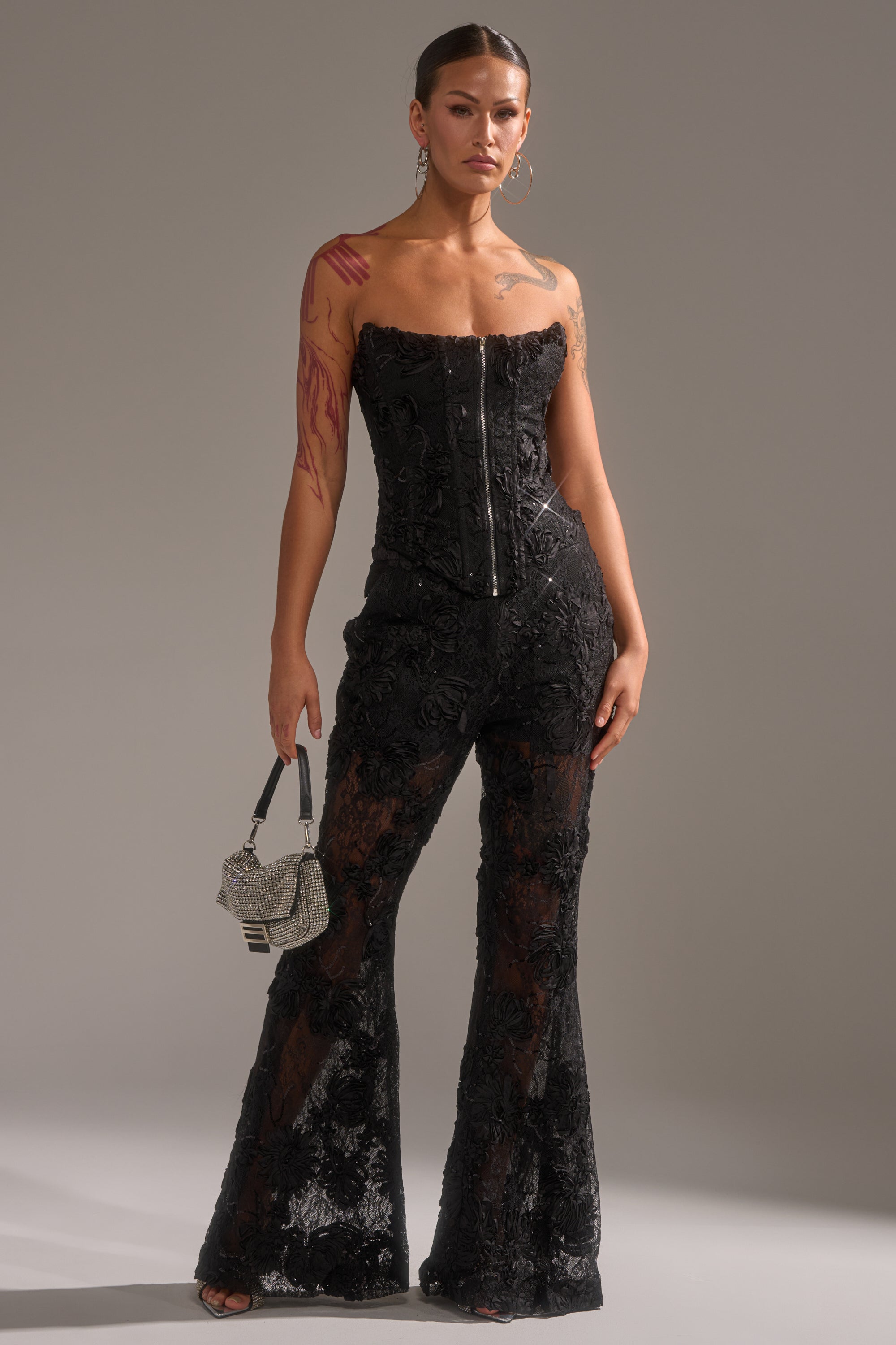 A person stands confidently in the LET IT GO LACE TROUSER—high-rise, flared pants with sheer embroidered details—paired with a strapless black corset top, silver bag, visible tattoos, earrings, and slicked-back hair.