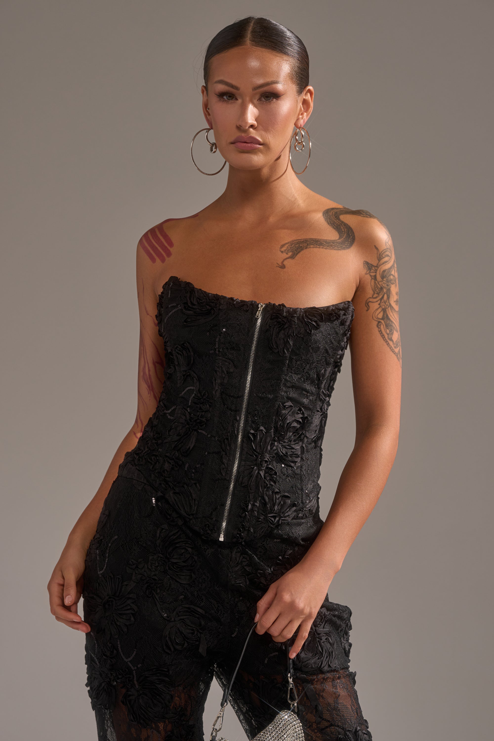 A tattooed woman wears the LET IT GO LACE CORSET—strapless with 3D textured detailing—paired with black pants. She holds a small silver handbag and has her hair sleekly pulled back, standing against a neutral backdrop.