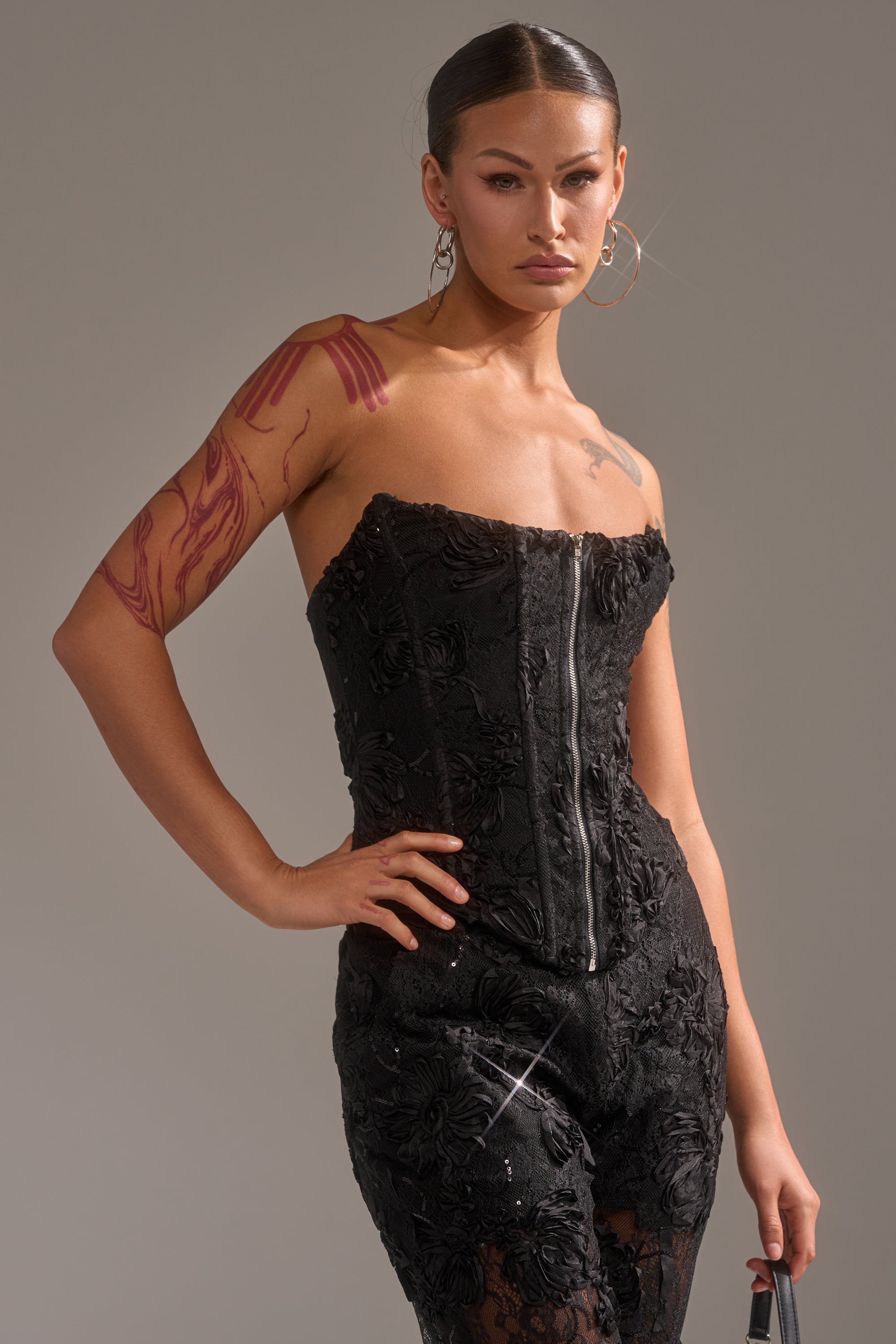 A person with hoop earrings and tattoos poses confidently in the LET IT GO LACE CORSET, which features 3D textured detailing and a front zipper, hand on hip against a neutral background.