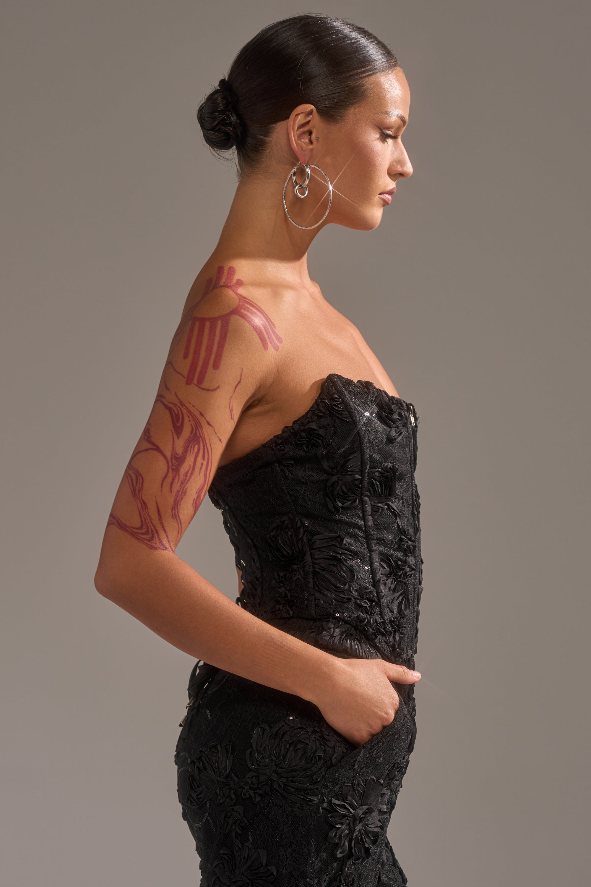 A woman in the LET IT GO LACE CORSET dress stands in profile with her hand in her pocket, wearing large hoop earrings, a sleek bun, and showing a red abstract upper arm tattoo against a plain gray background.
