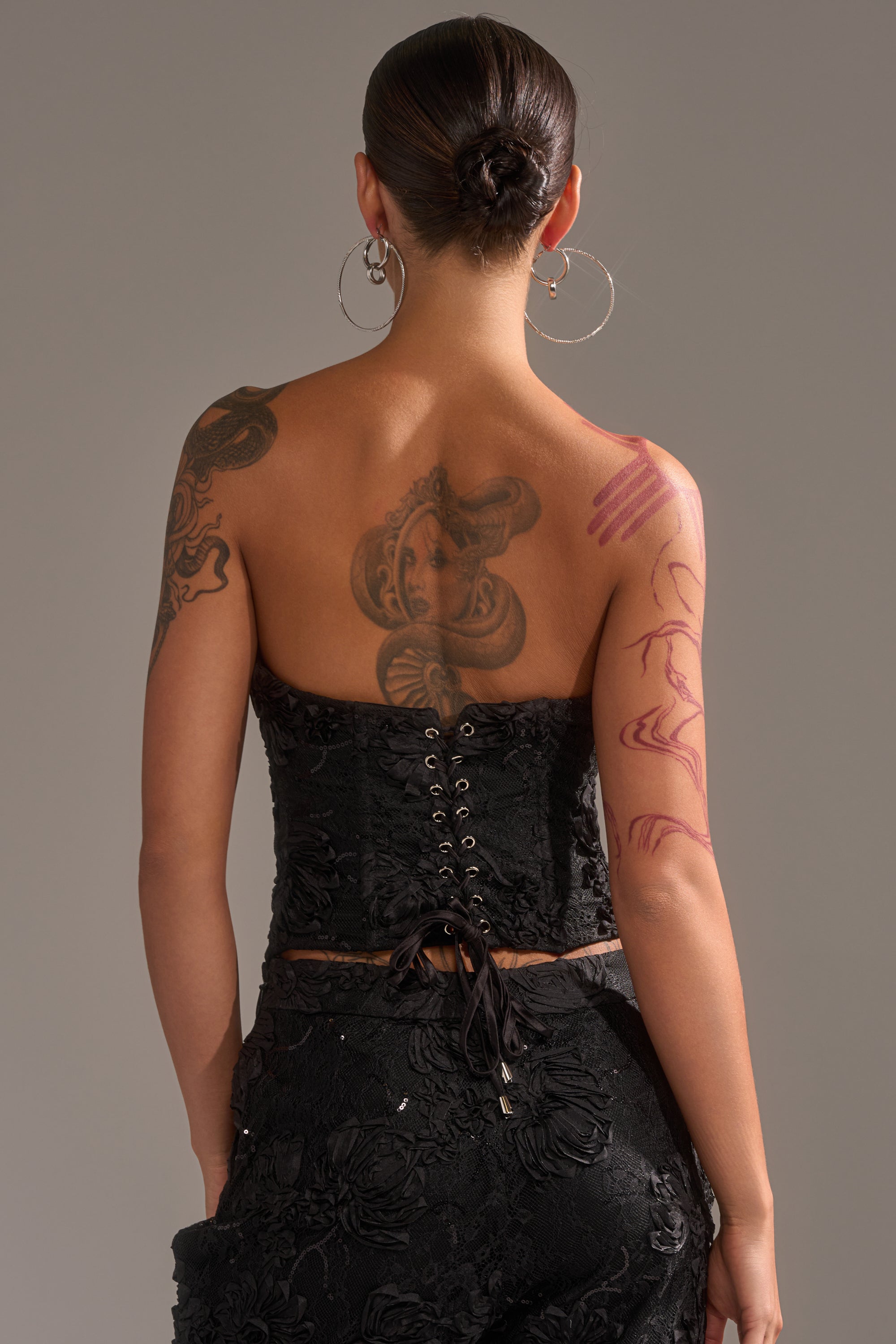 A person with large hoop earrings and back and arm tattoos is shown from behind in a sleek bun, wearing the strapless LET IT GO LACE CORSET with 3D textured detailing and lace-up accents, paired with a matching skirt.