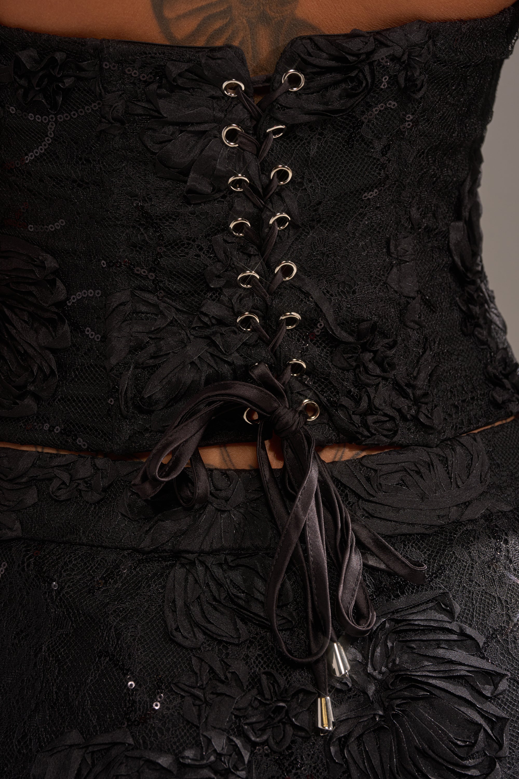 Close-up of the back of the LET IT GO LACE CORSET in black floral mesh lace, featuring delicate patterns, black ribbon lacing through metal grommets, and a neat bow at the waist. Lower back and part of a tattoo are visible.