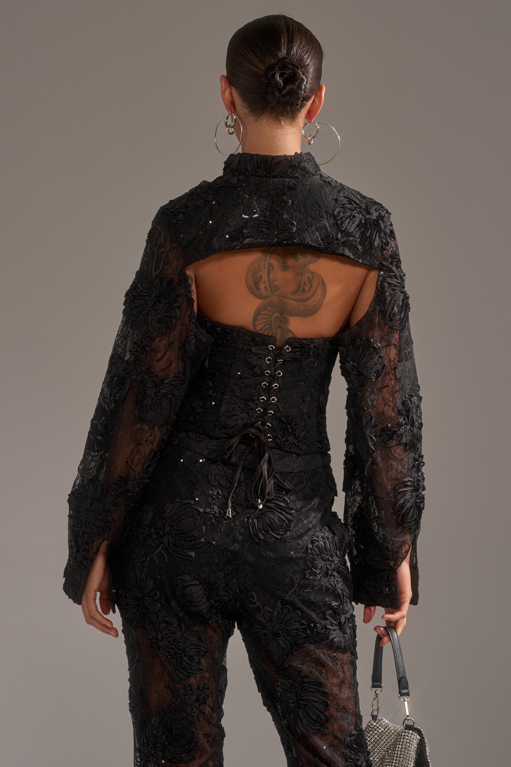 A person with their hair in a bun wears the LET IT GO LACE JACKET, featuring black textured fabric, an open back, and lace-up detail. A tattoo is visible on their upper back as they carry a small handbag and wear large hoop earrings.