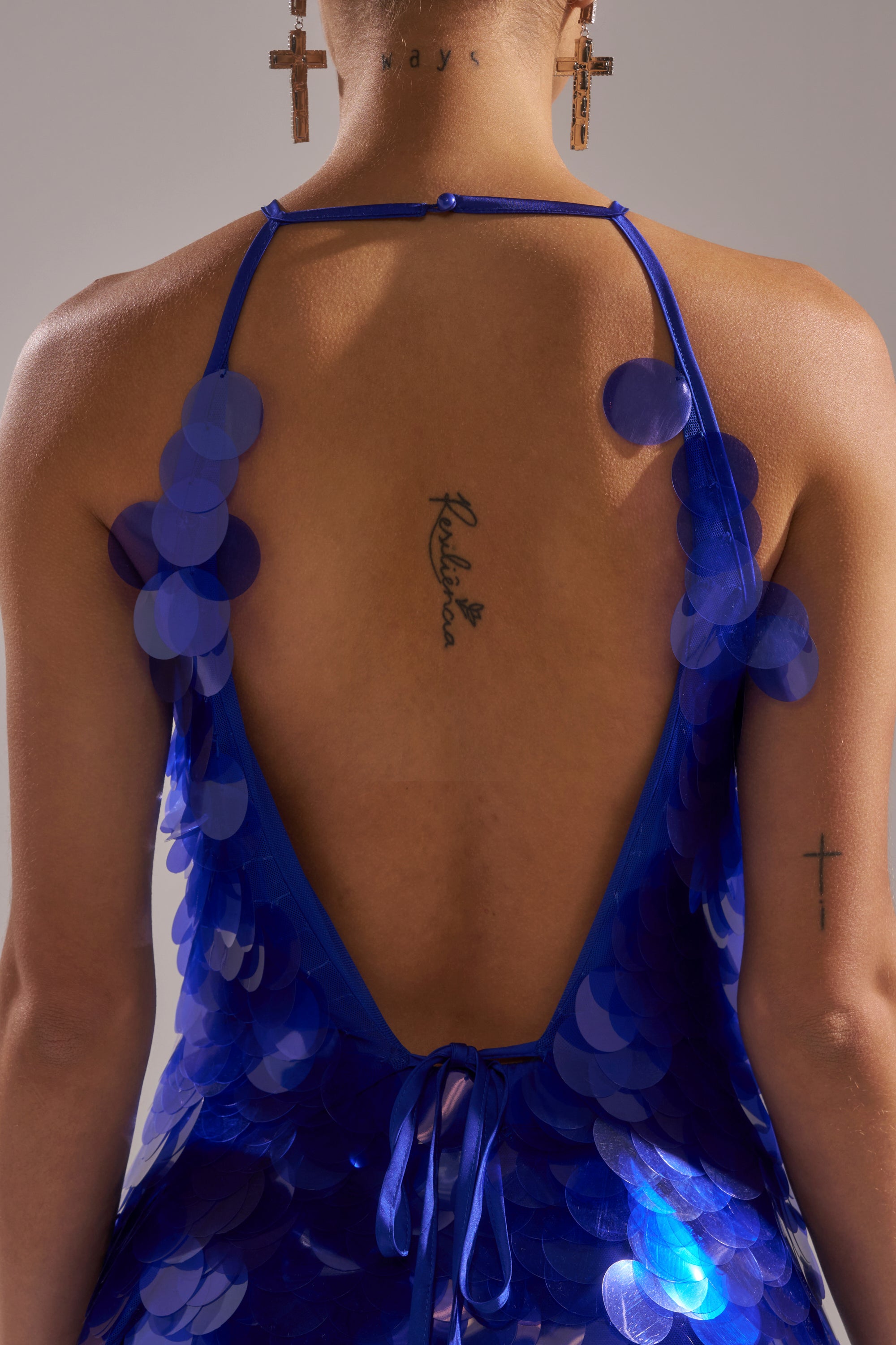 A person wears the DRAMA SEQUIN MINI DRESS in blue, featuring a low open back that reveals tattoos on their upper back and arm, paired with cross-shaped earrings.