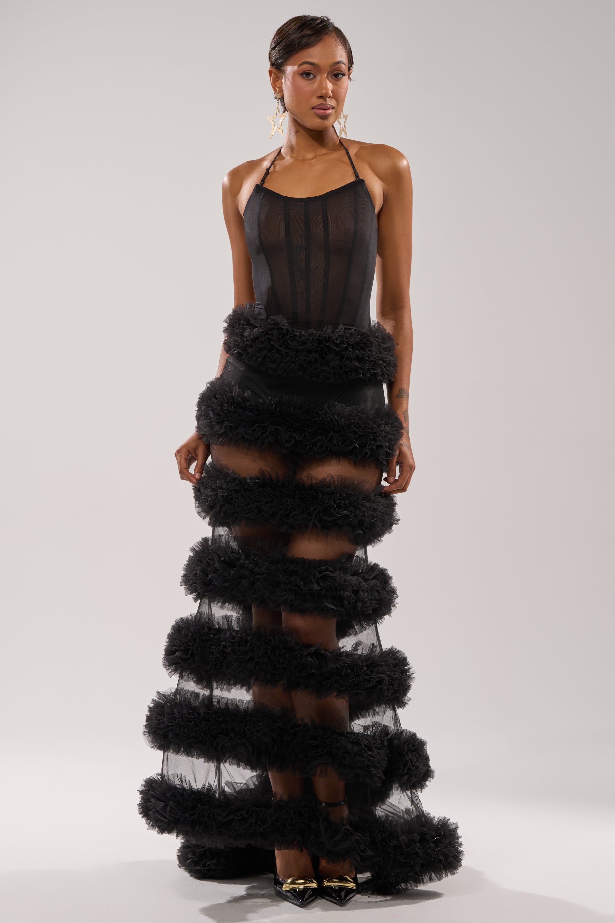 She wears the QUEEN OF EVERYTHING MAXI DRESS, a black sheer tulle maxi with horizontal ruffles from waist to hem, styled with gold pointed shoes and dangling earrings, posing against a plain light background. 0
