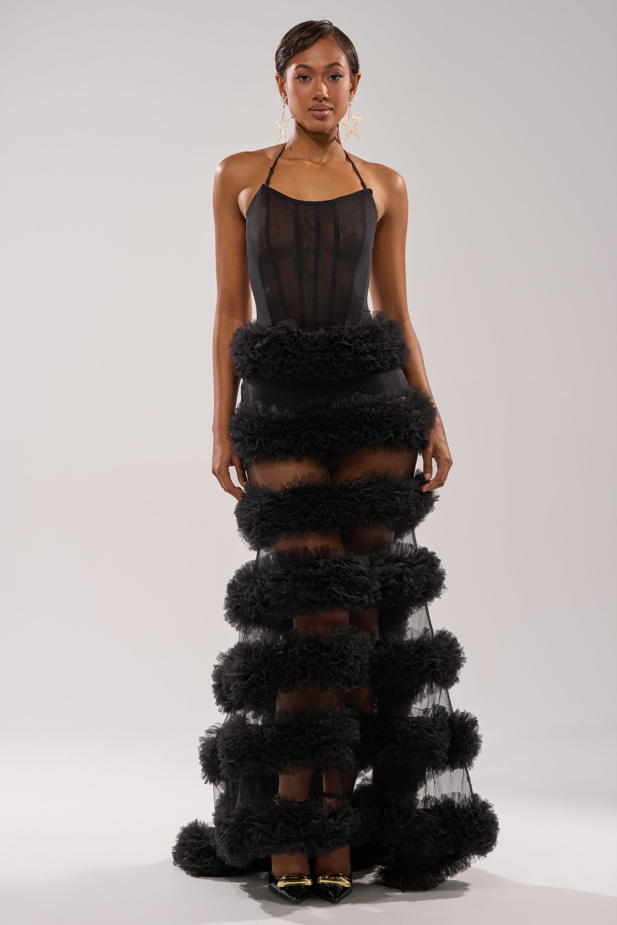 A woman faces forward in the QUEEN OF EVERYTHING MAXI DRESS, a sheer black gown with a corset bodice and ruffled bands down the skirt. She pairs it with statement star earrings and gold pointed-toe heels against a light background. 1