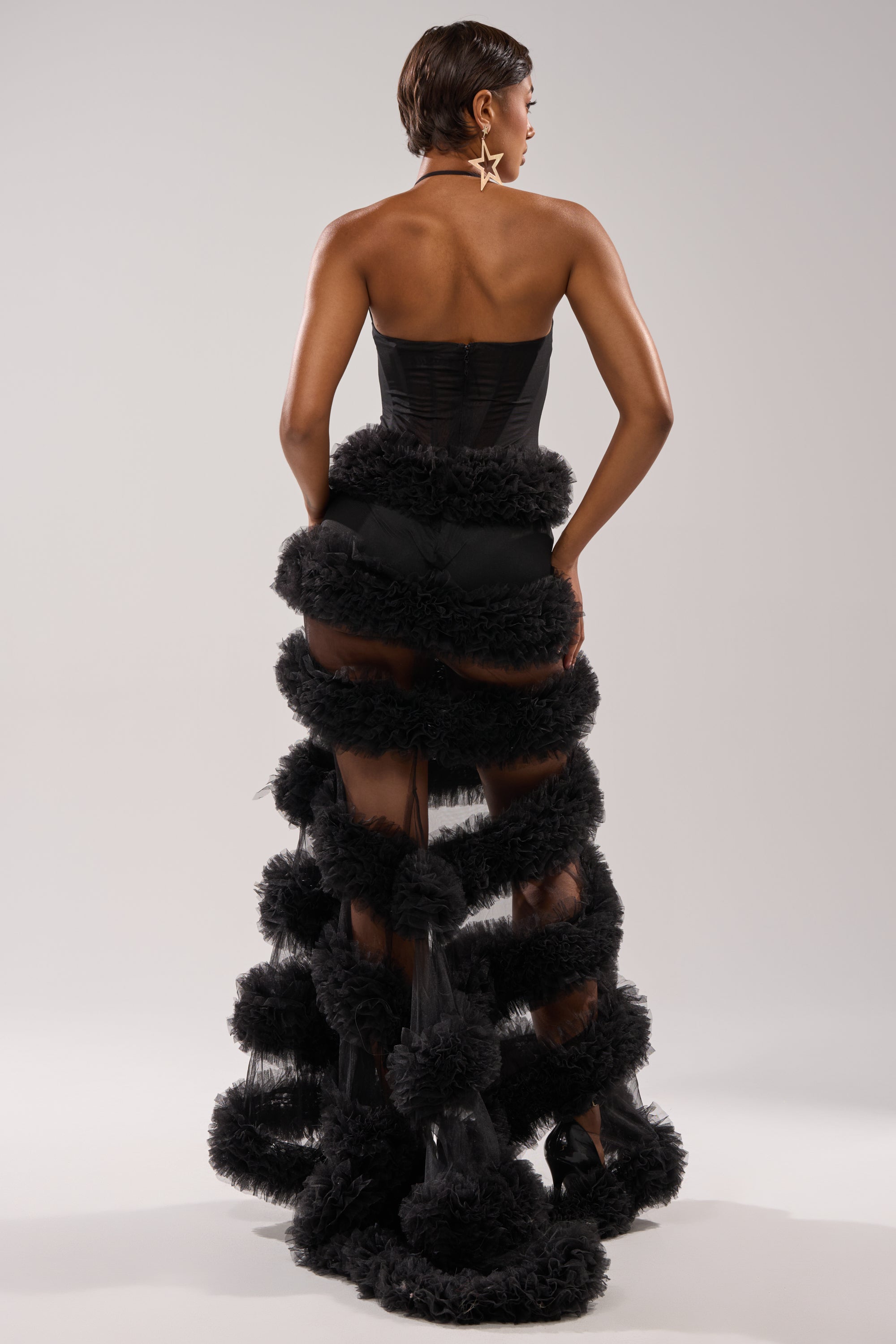 A woman with short hair and star-shaped earrings stands facing away, wearing the QUEEN OF EVERYTHING MAXI DRESS—a strapless black sheer tulle dress with a corset-style bodice and layered ruffles—set against a plain light gray background. 3