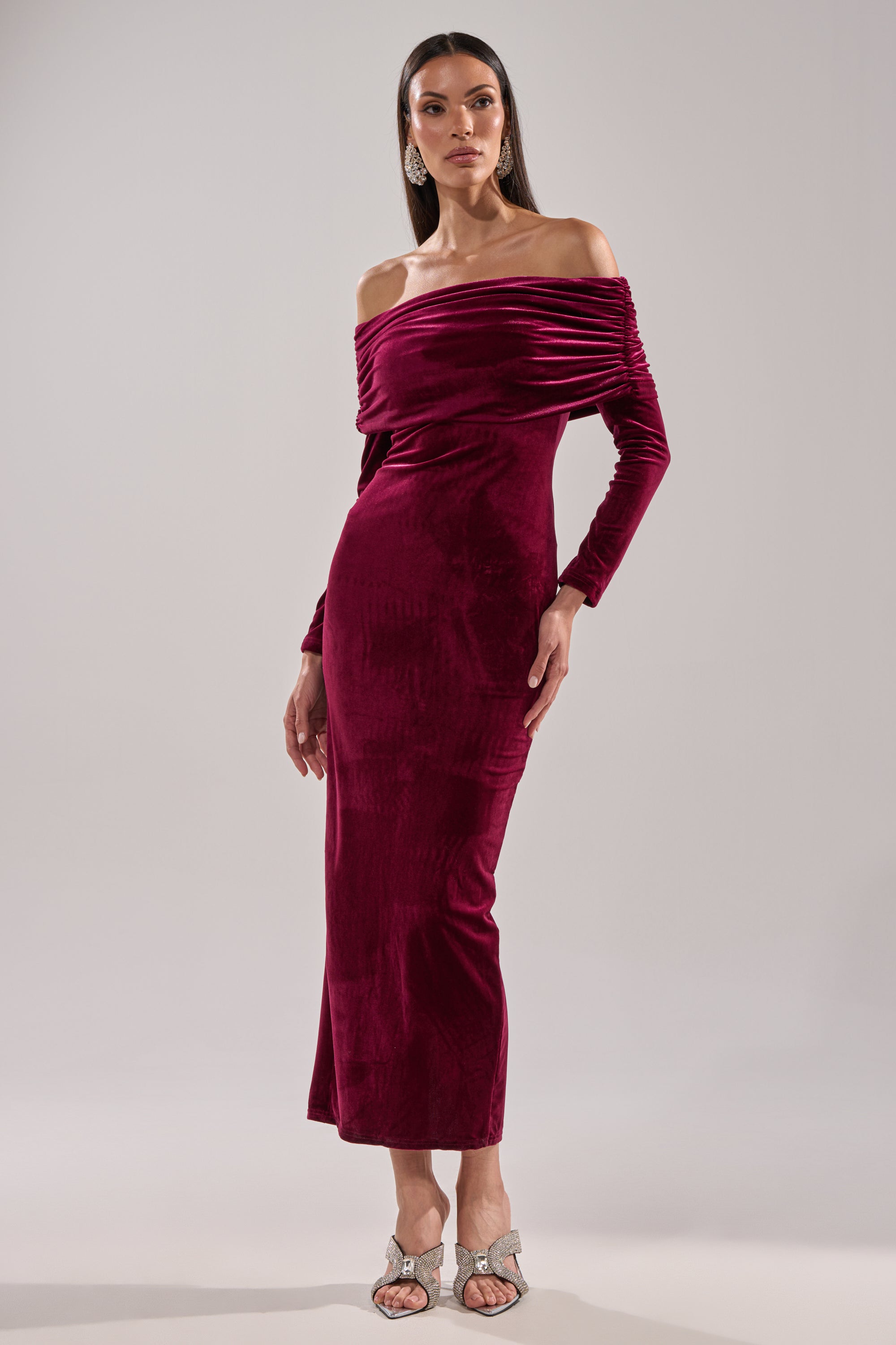 A woman wears the IN THE AIR VELVET MAXI DRESS, a long burgundy off-the-shoulder velvet maxi with long sleeves, paired with silver high-heeled sandals, set against a plain light background.