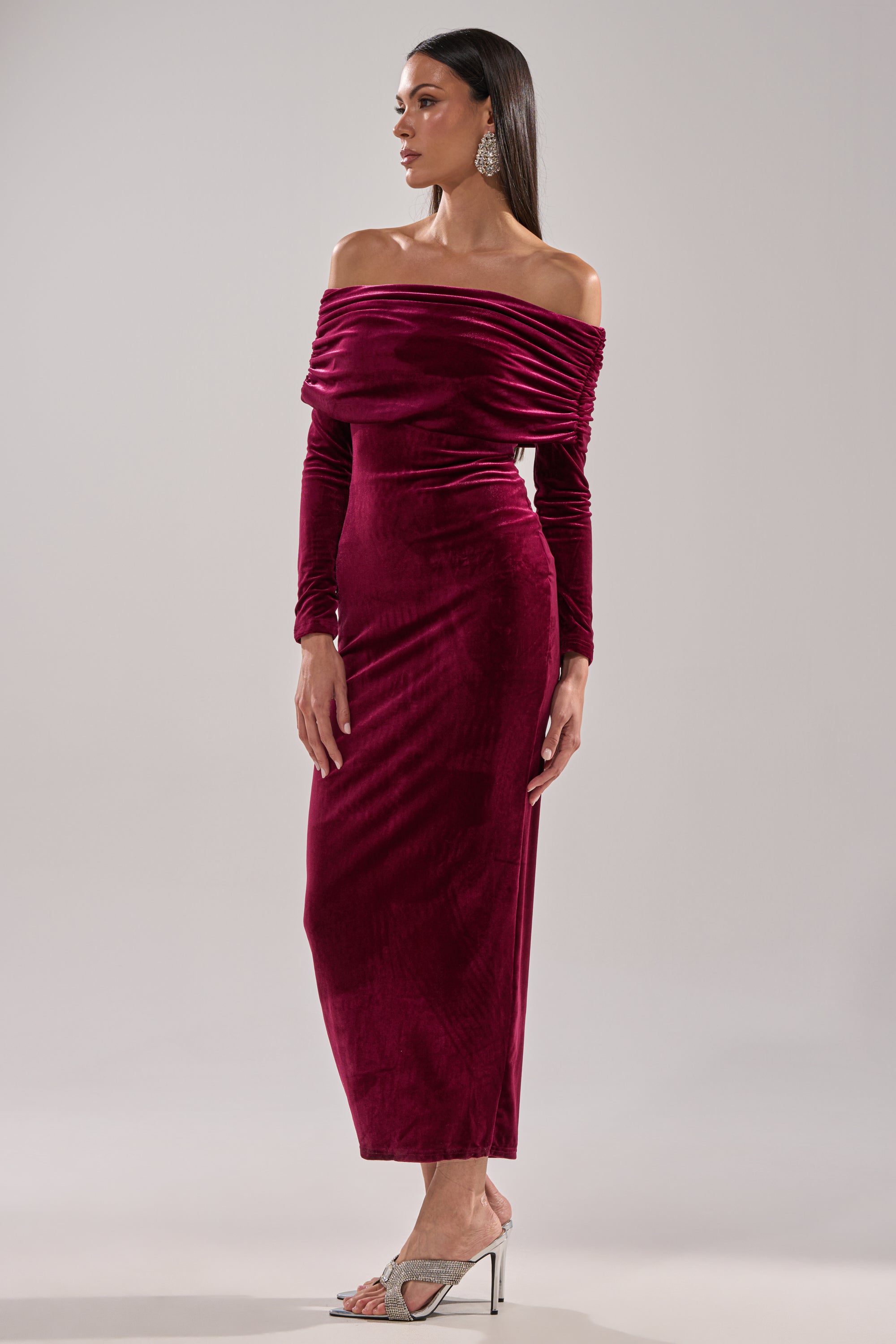 A woman models the IN THE AIR VELVET MAXI DRESS in deep red, featuring an off-the-shoulder neckline and long sleeves, paired with silver high-heeled sandals, standing against a plain background and looking to her left.