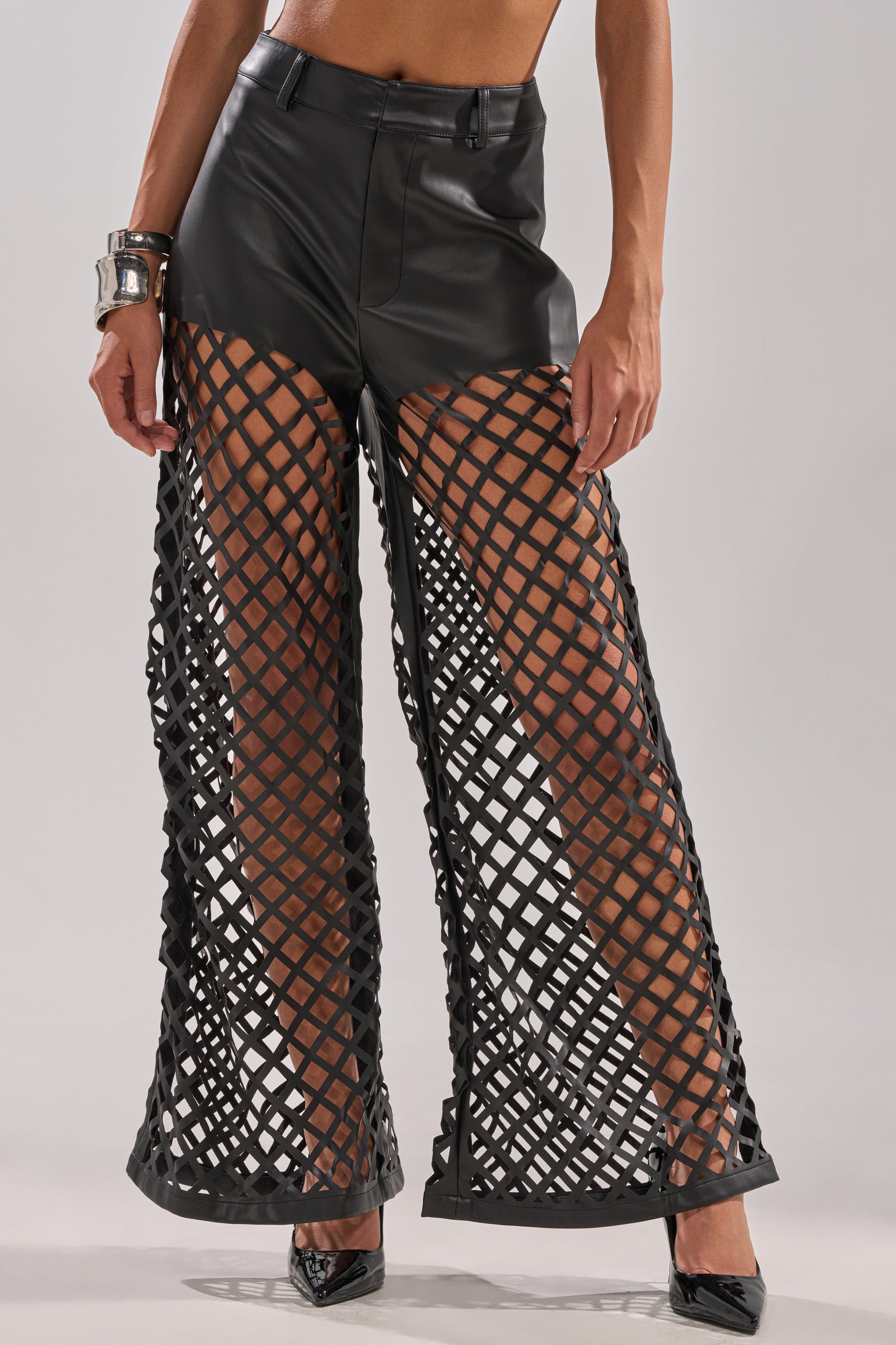 Wearing the OPEN UP FAUX LEATHER TROUSER featuring a flared-leg silhouette and bold lattice cut-outs, styled with black heels and a chunky silver bracelet for an edgy look. 1
