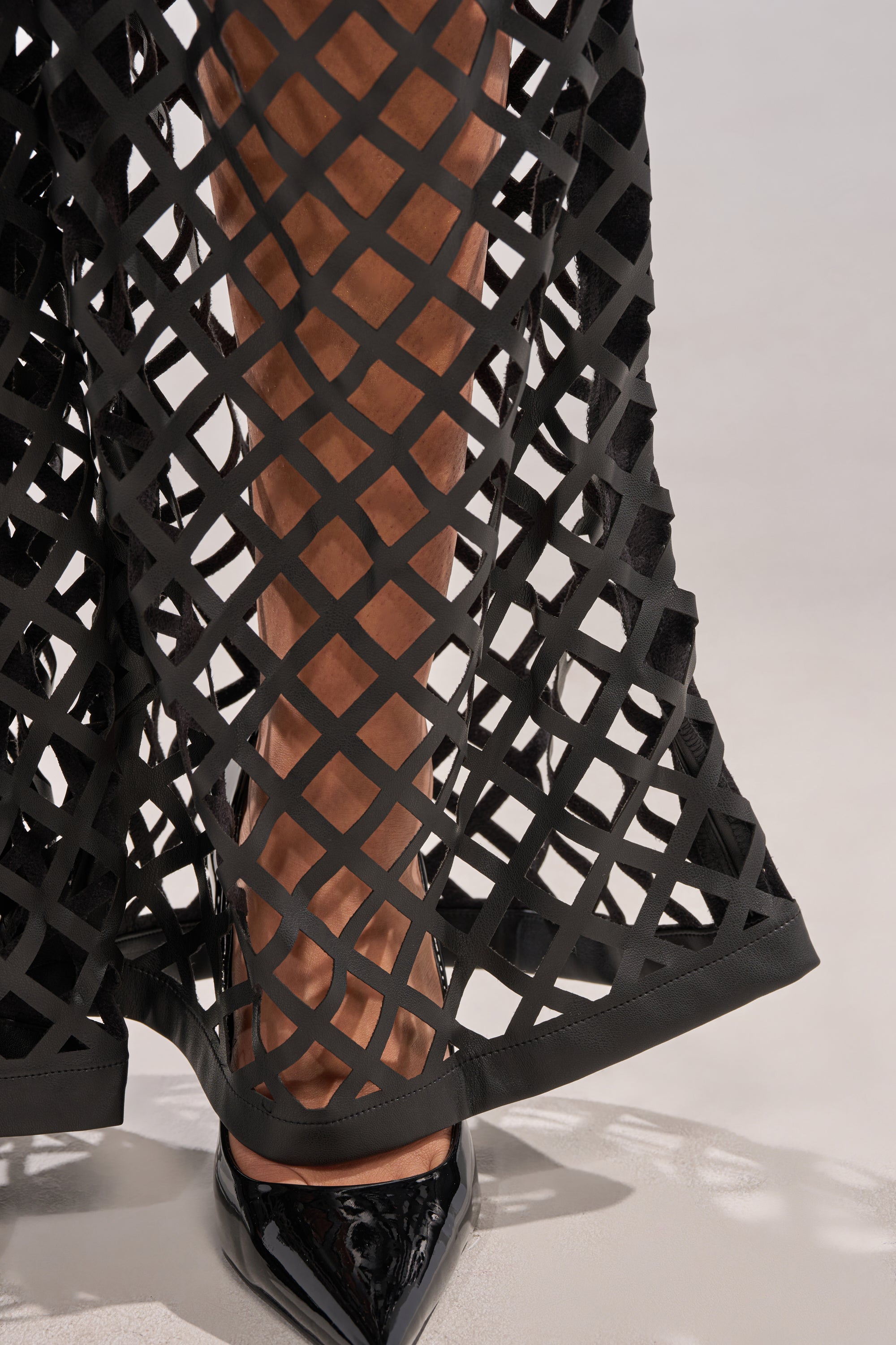 A close-up of a person wearing the OPEN UP FAUX LEATHER TROUSER, featuring black lattice-patterned sheer panels and cut-out details, reveals their legs underneath as they stand in shiny black pointed-toe heels. 8
