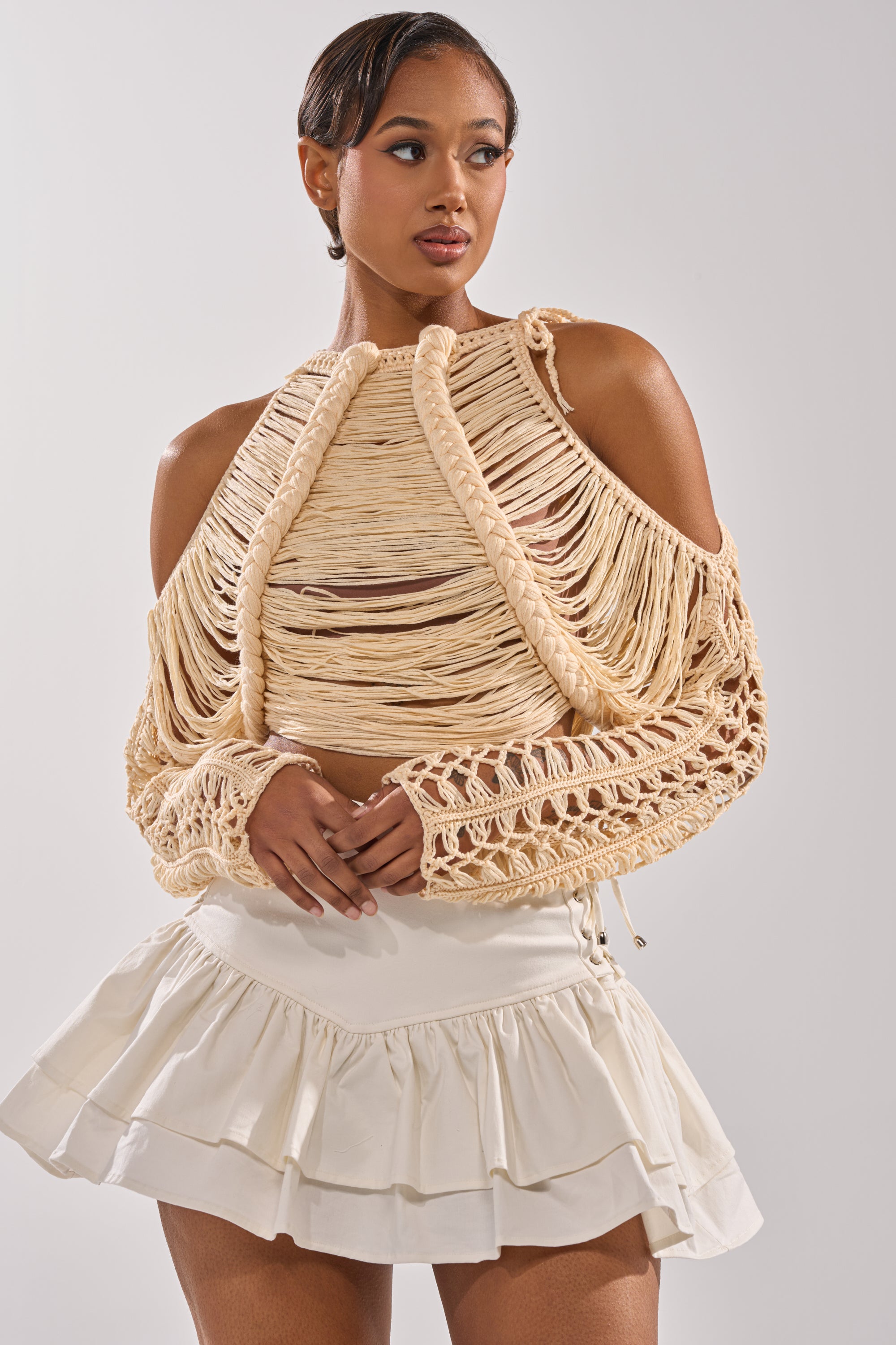 A woman poses against a plain light background in the CATCHING FEELING FRINGE SWEATER—a beige macramé knit with open shoulders and long sleeves—paired with a white ruffled mini skirt. 0