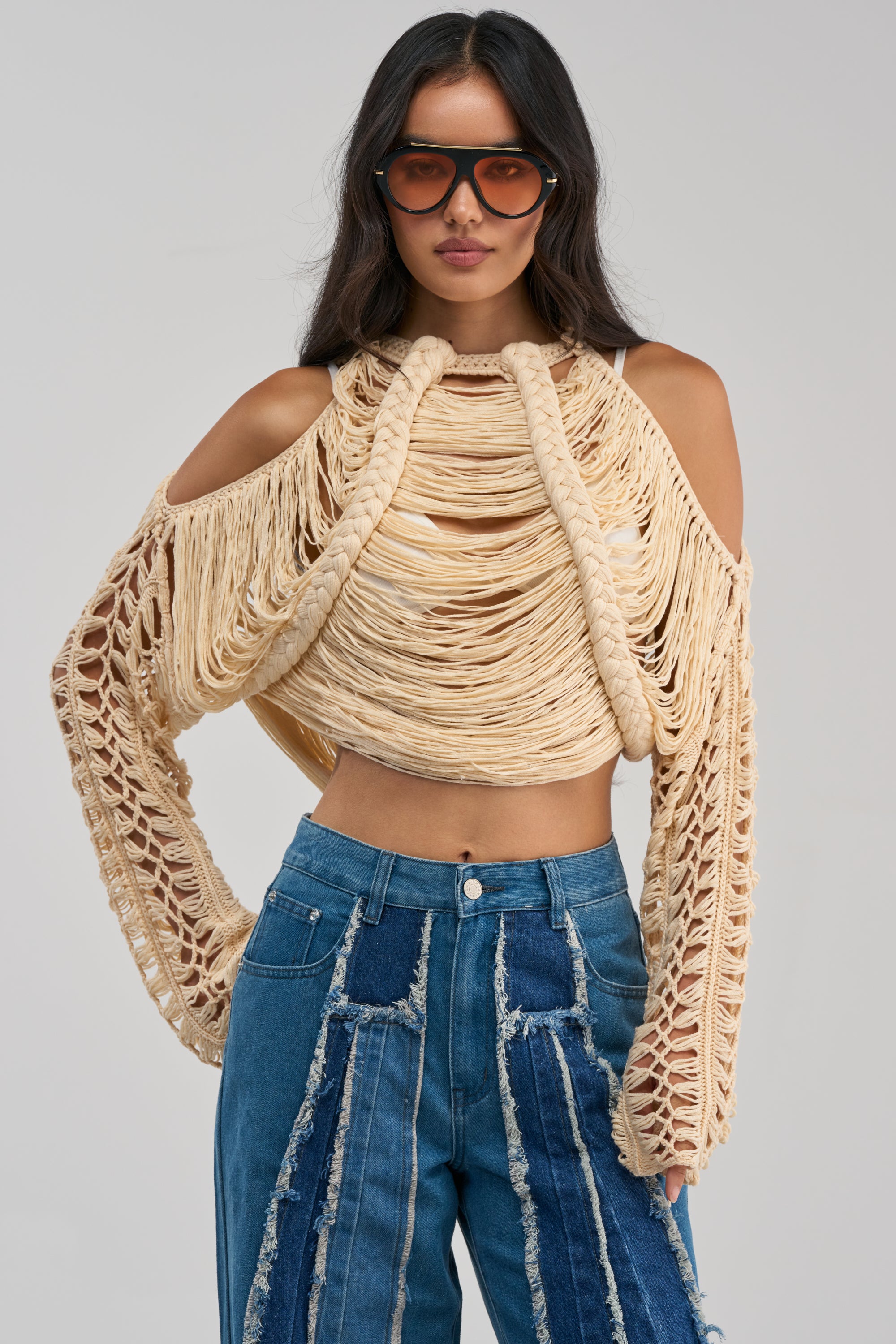 A woman stands facing forward against a plain background, wearing large sunglasses, blue patchwork denim jeans, and the CATCHING FEELING FRINGE SWEATER—a beige open-weave sweater with cut-out shoulders featuring a rope-like design.