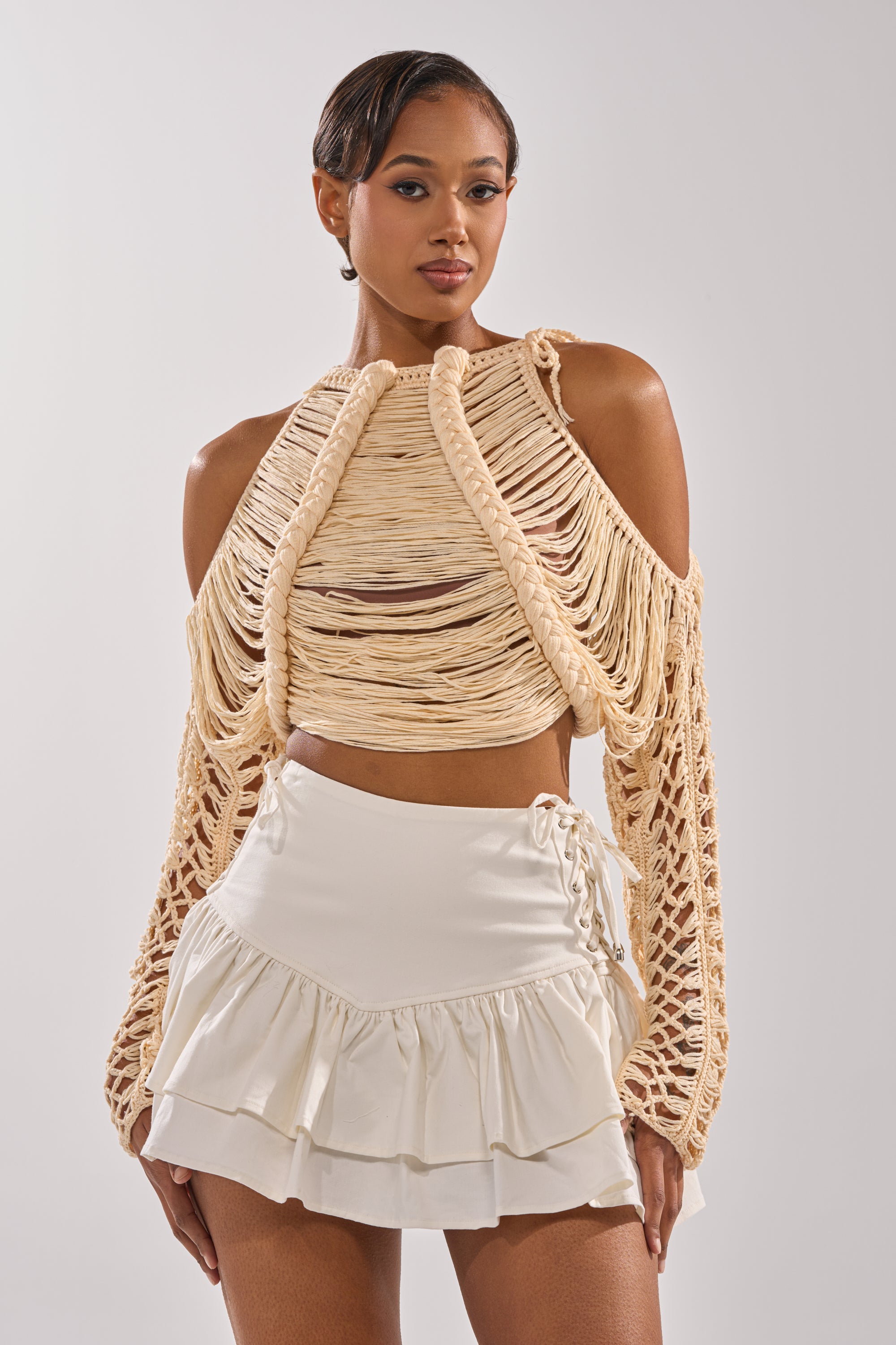 A woman with short, sleek hair and a neutral expression poses confidently against a plain background, wearing the beige CATCHING FEELING FRINGE SWEATER with draped sleeves and a cream ruffled mini skirt. 1