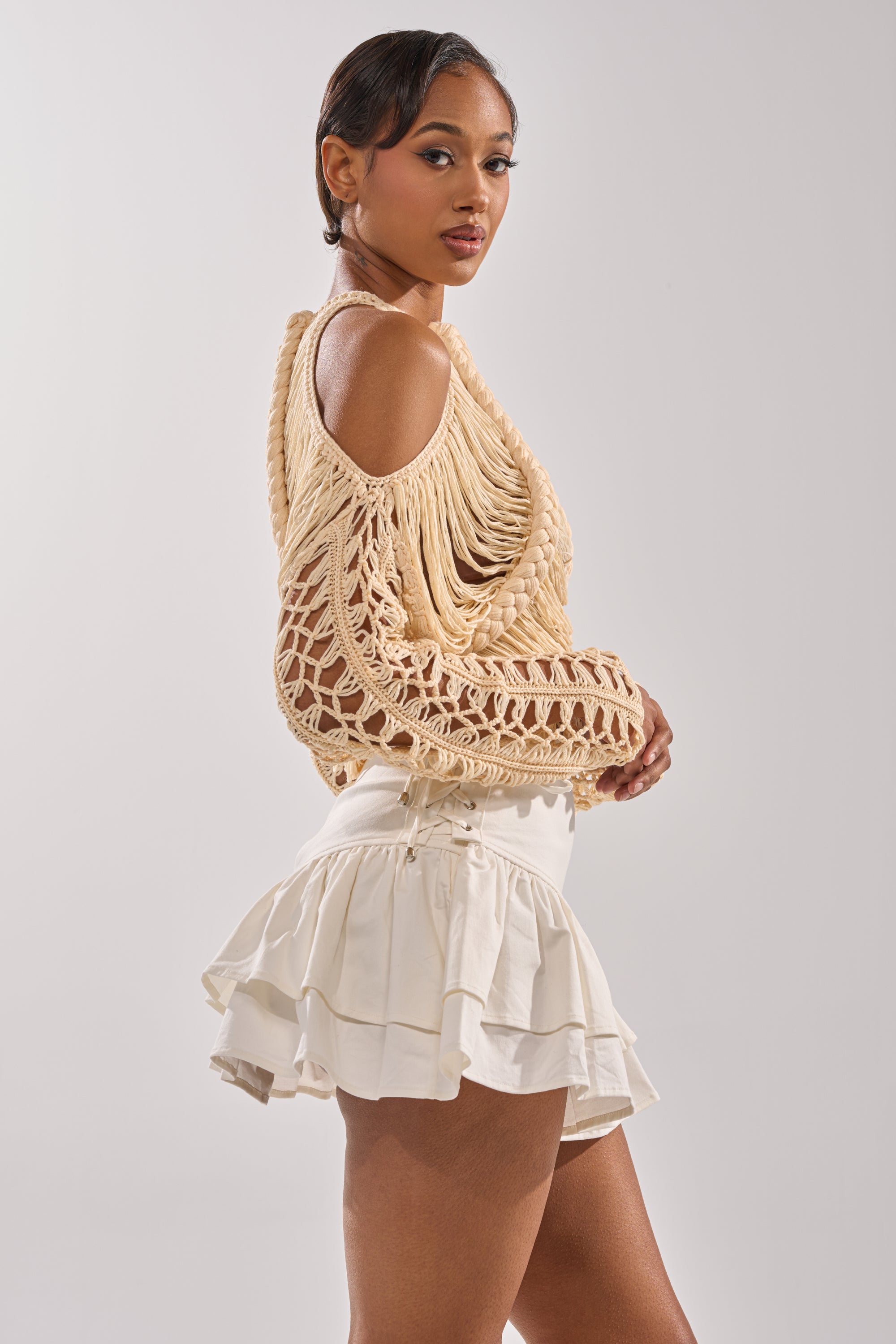 A woman poses sideways in the CATCHING FEELING FRINGE SWEATER, a beige knit macramé off-shoulder style, paired with a white layered ruffled mini skirt, her hair styled back against a plain light background. 2