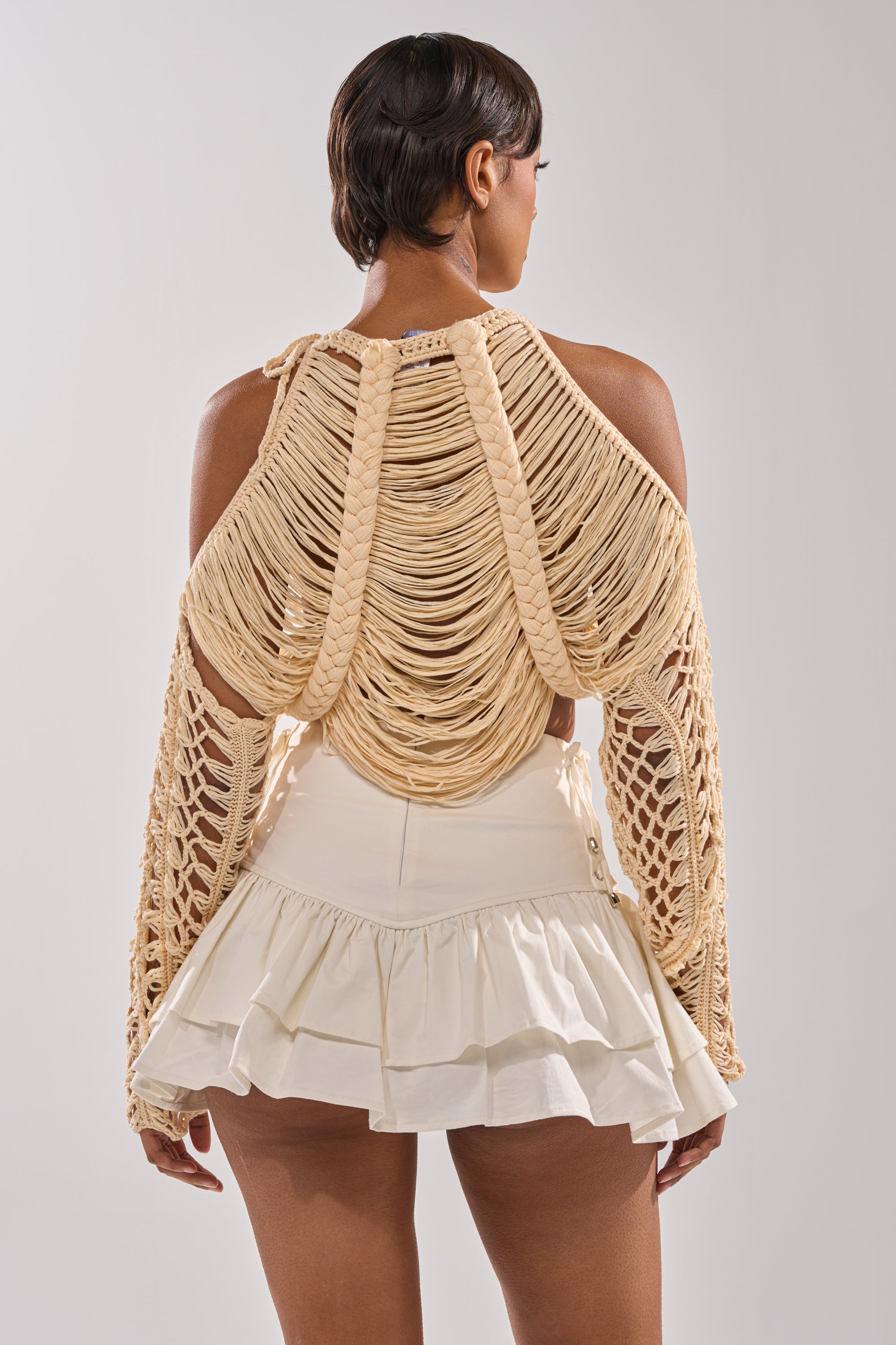 A person stands with their back to the camera, wearing the CATCHING FEELING FRINGE SWEATER—cream-colored, knit with thick braided strands and open sleeves—paired with a high-waisted, ruffled white mini skirt. 3