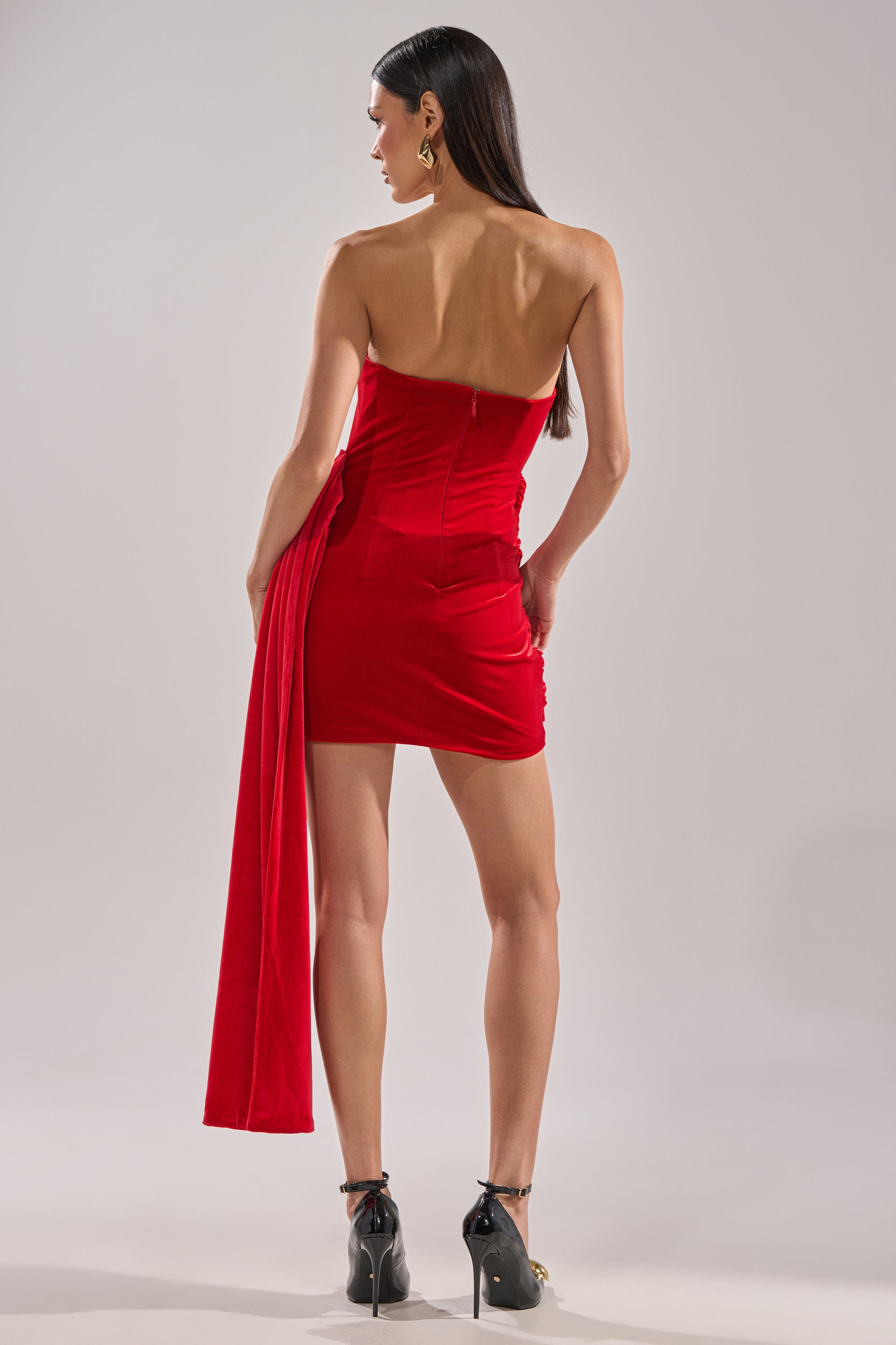 A woman with long dark hair faces away, wearing the ON FIRE VELVET MINI DRESS—a strapless red velvet mini with asymmetric draping—and black high-heeled sandals. The background is a plain, light gray.
