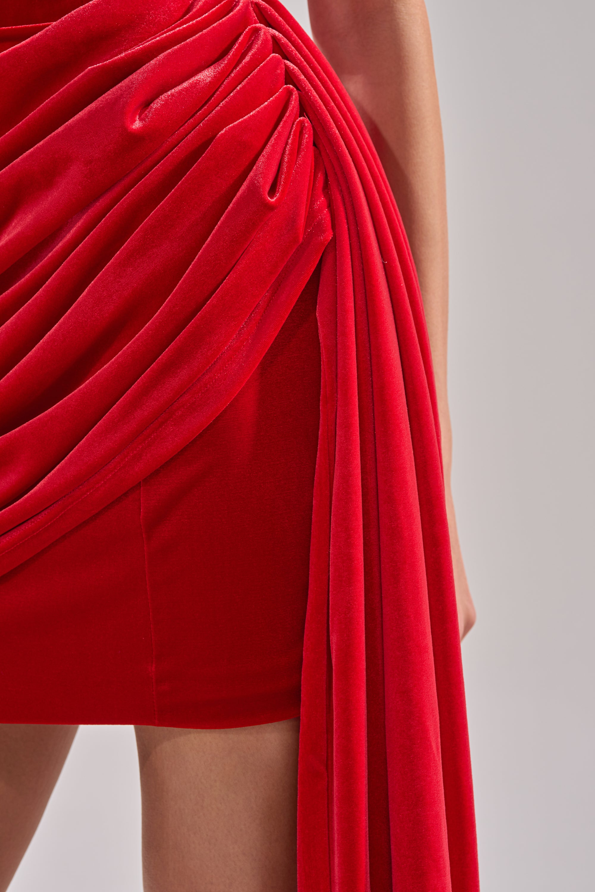 Close-up of a person wearing the ON FIRE VELVET MINI DRESS—a vibrant red mini dress with elegant draping—revealing one arm and part of the legs against a neutral background.
