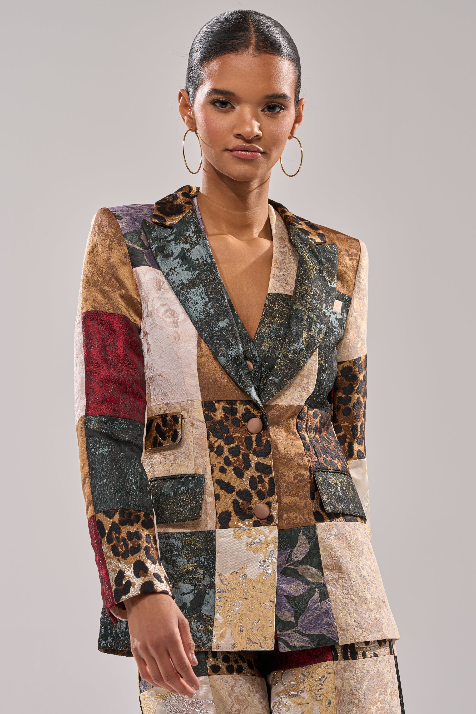 Woman in the MADE YOU LOOK PATCHWORK BLAZER, a tailored jacket with floral, leopard print, and metallic patchwork. She has slicked-back hair and finishes her bold look with large hoop earrings.