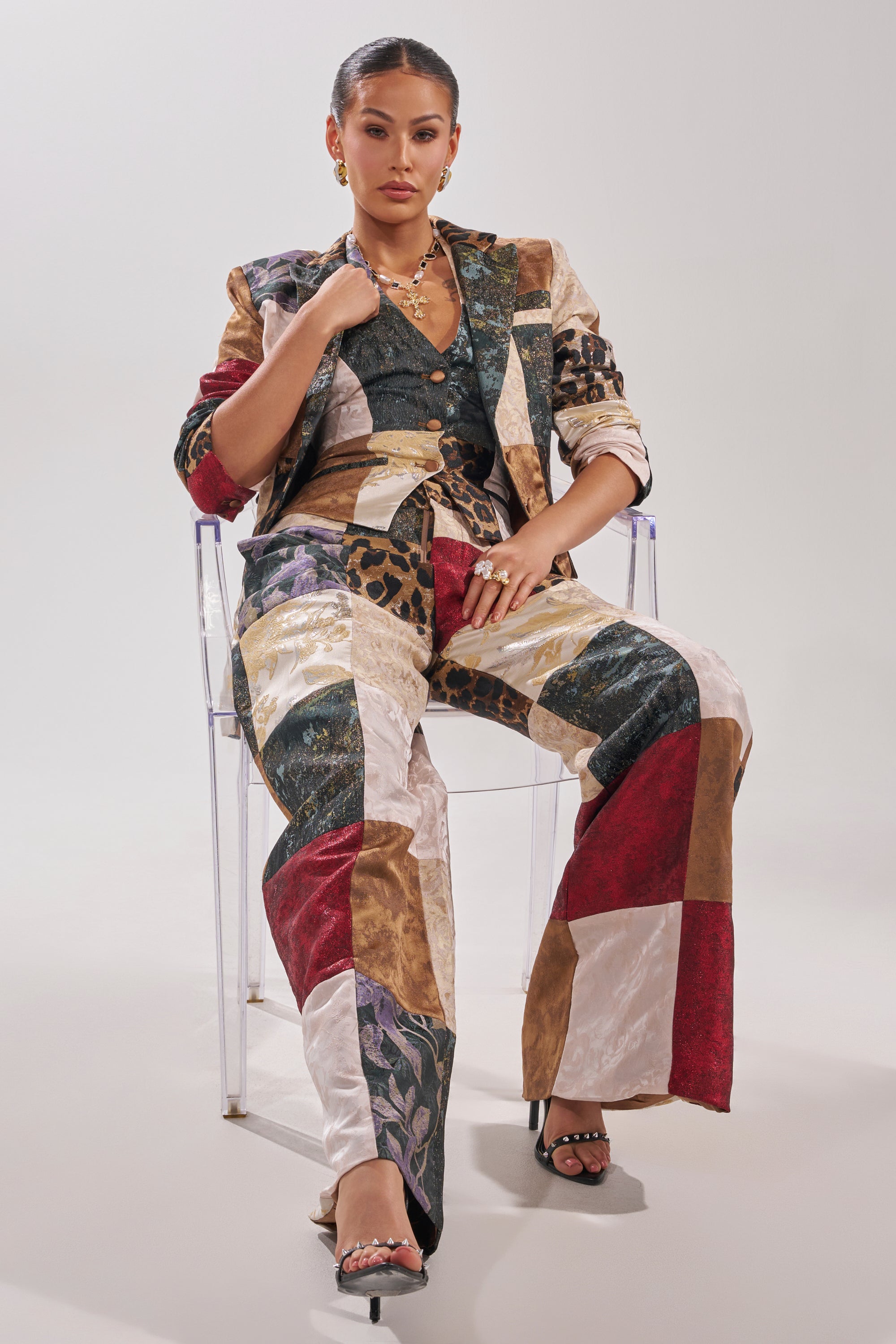 A woman sits confidently on a clear chair, wearing the MADE YOU LOOK PATCHWORK BLAZER featuring bold animal print and floral designs. Her hair is pulled back, styled with hoop earrings and layered necklaces.